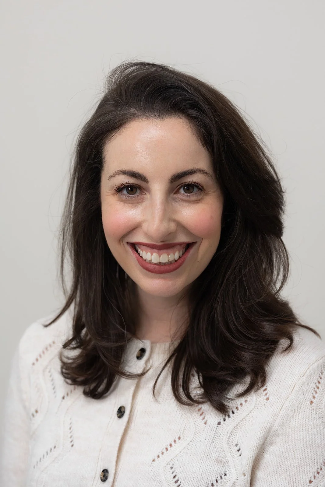 A woman with long brown hair smiling, wearing a cream-colored sweater with textured patterns, standing in front of a brick wall with sunlight.
