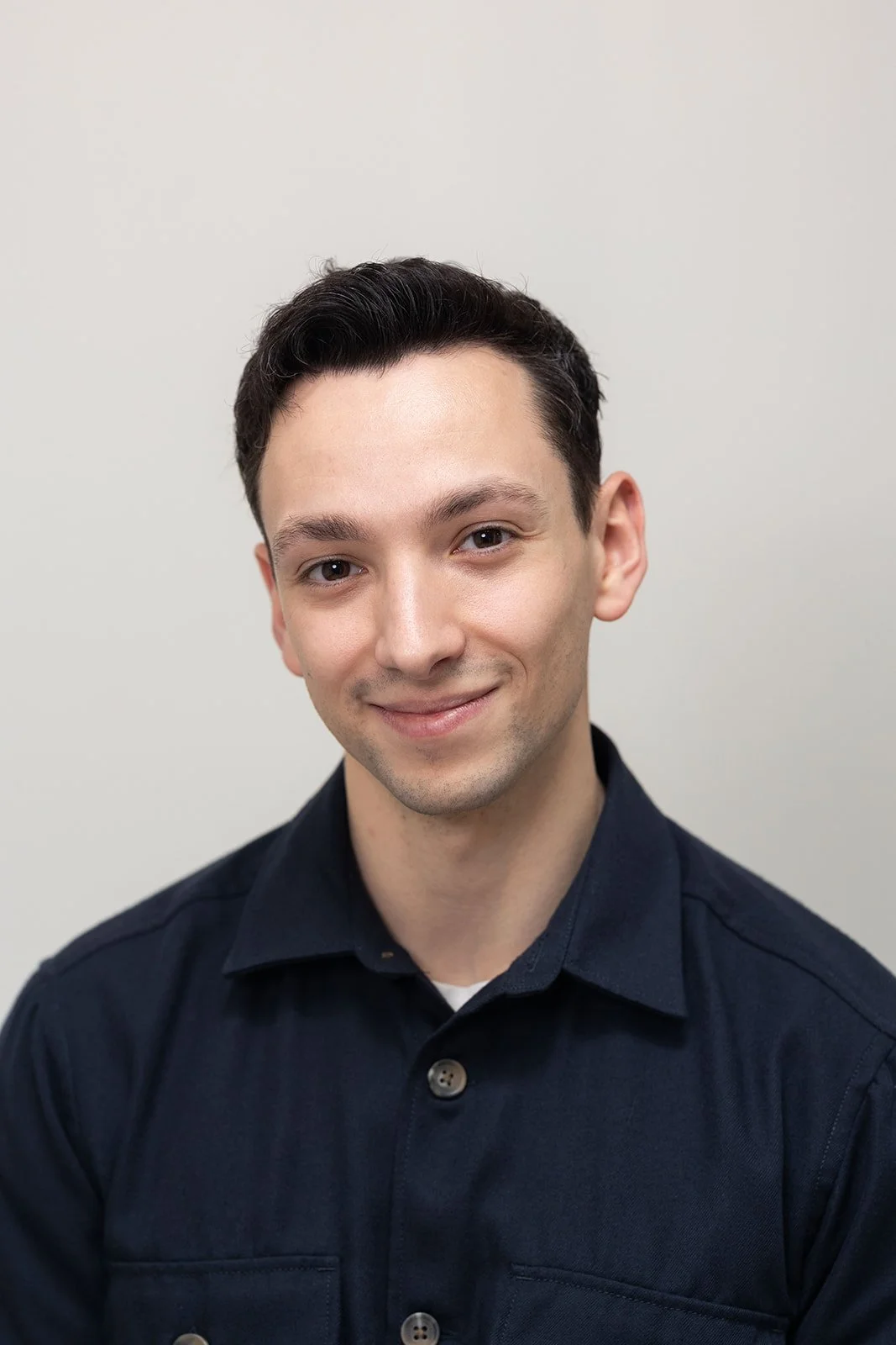 A young man smiling, wearing a maroon polo shirt and dark jeans, standing against a light blue background.