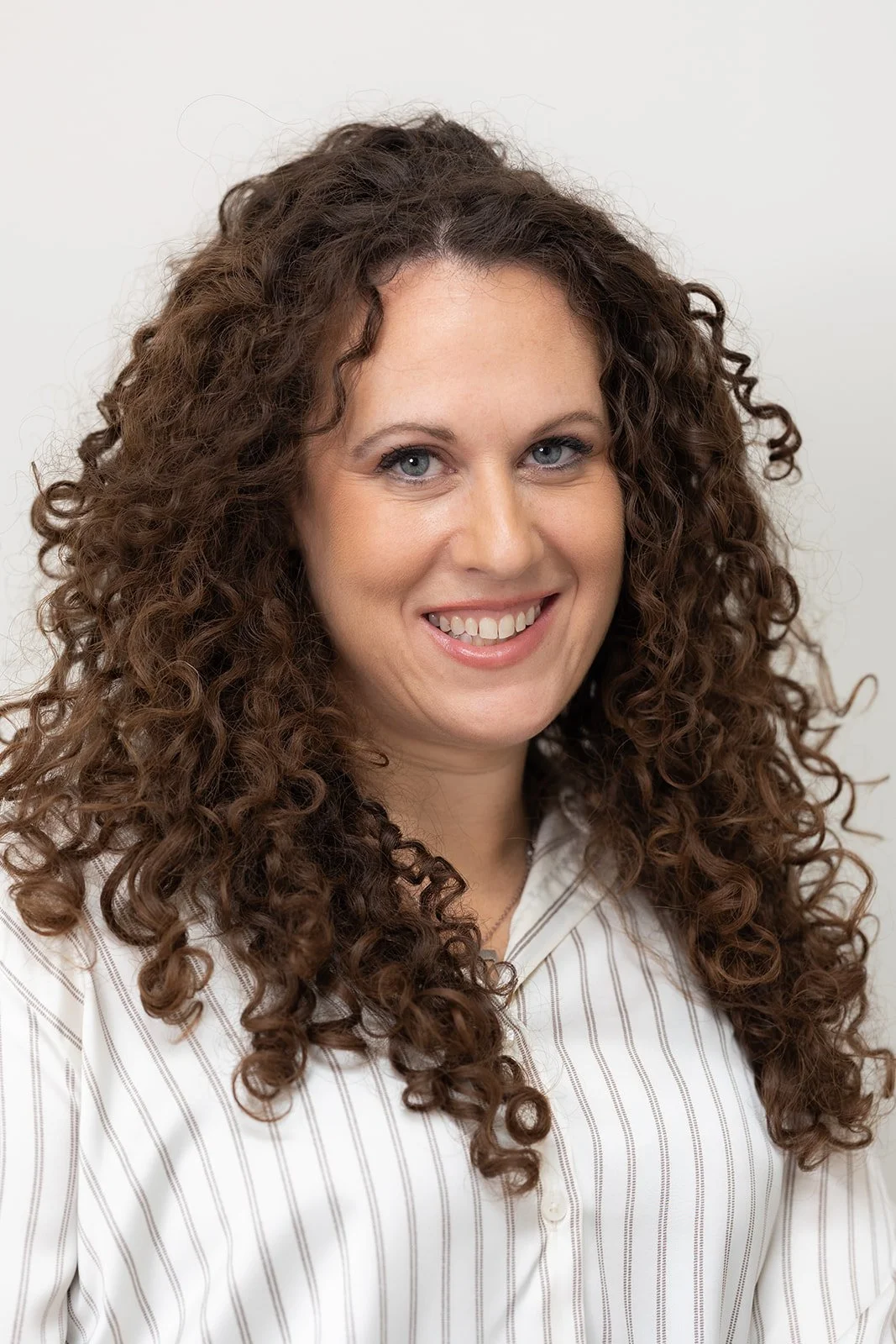 A woman with curly brown hair and hoop earrings, wearing a white lab coat, smiling in a professional setting.