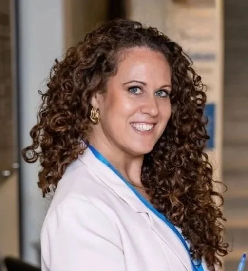 A woman with curly brown hair and hoop earrings, wearing a white lab coat, smiling in a professional setting.