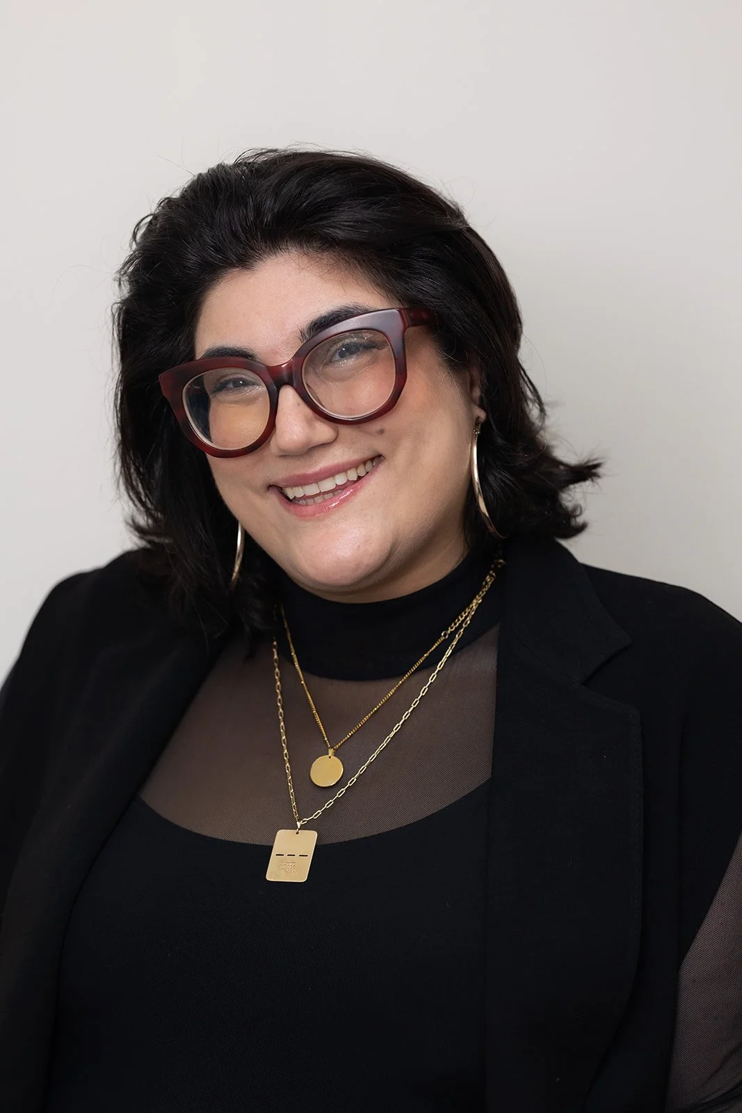 A smiling young woman wearing glasses and a light pink t-shirt with colorful text that reads 'Your Feelings Are Valid' in an indoor office or studio setting.