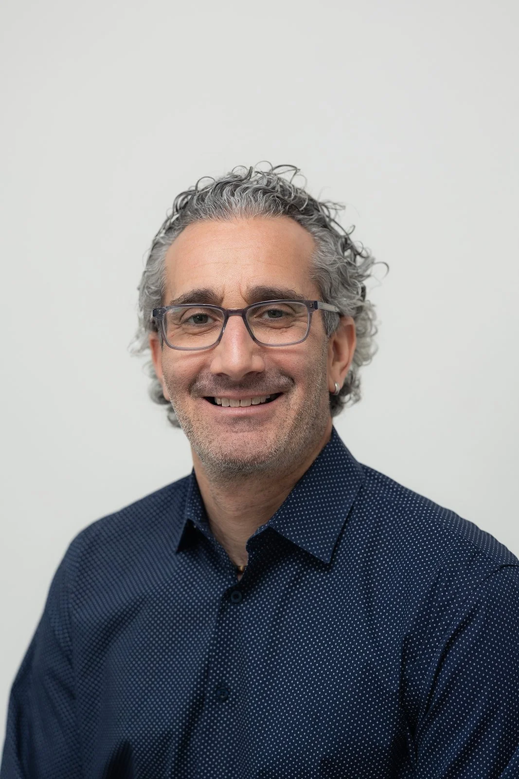 Close-up portrait of a smiling man with curly gray hair, glasses, and a green shirt against a dark background.