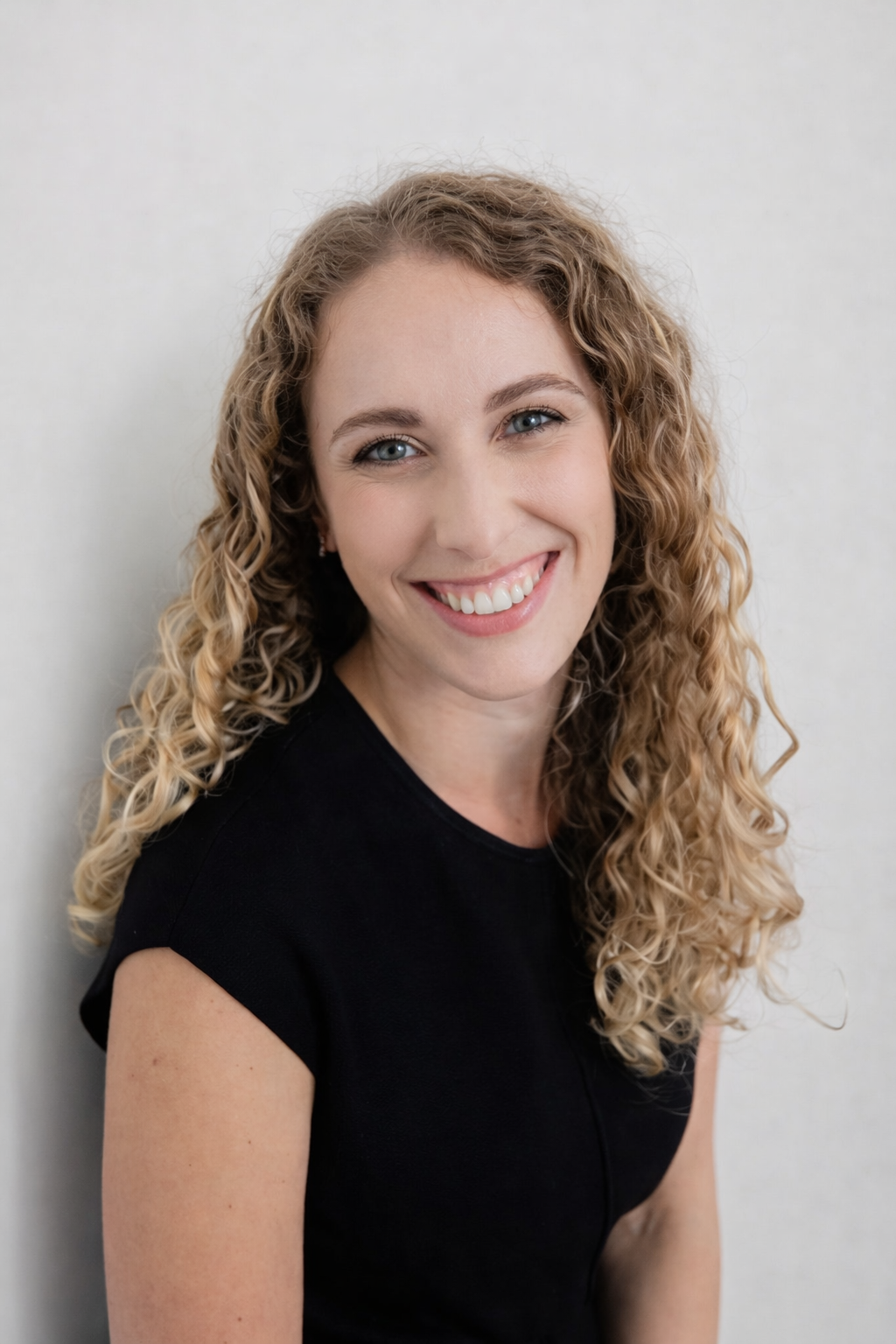 A young woman with curly blonde hair and a bright smile, wearing a black top, standing against a plain light-colored wall.
