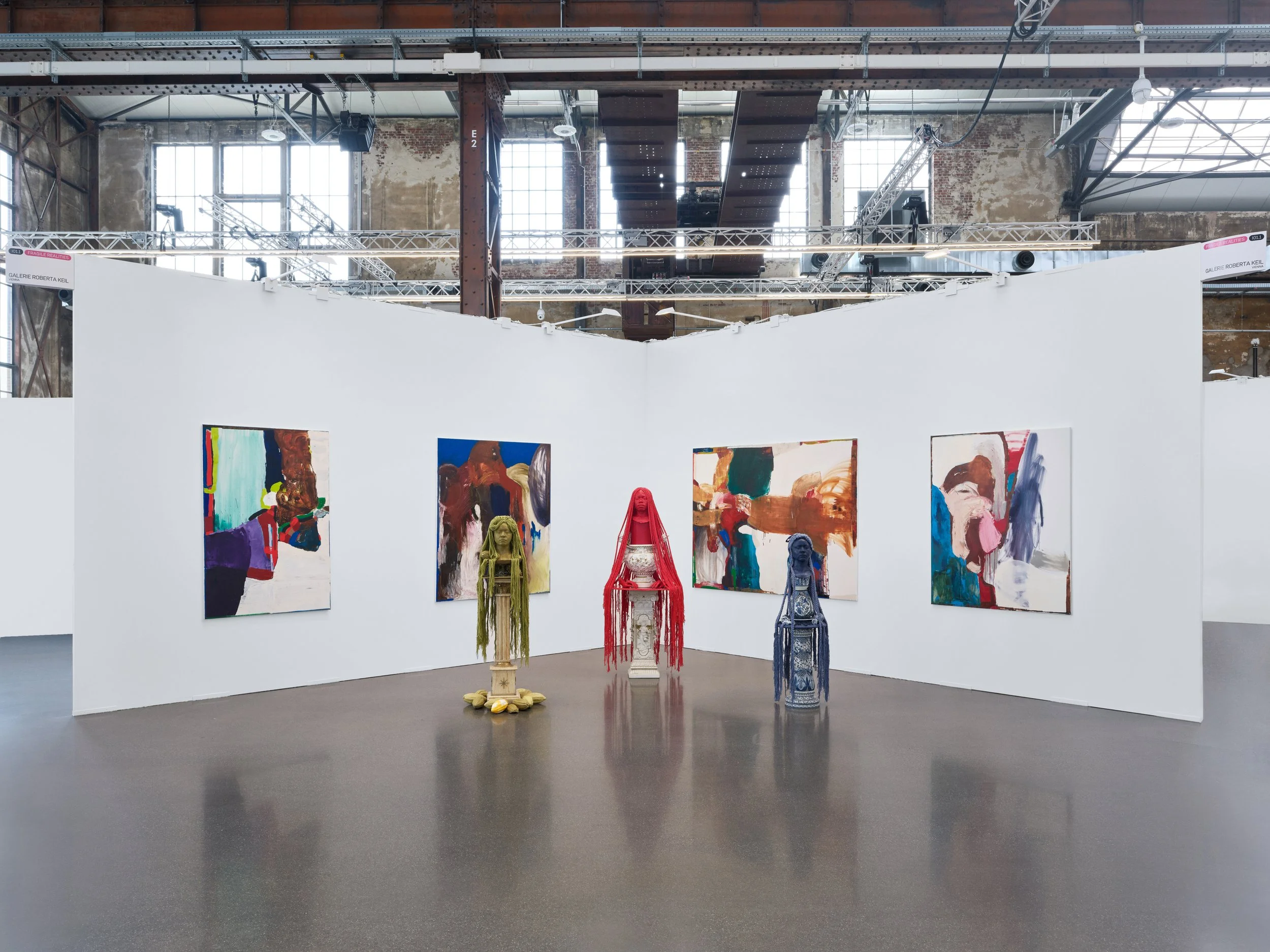 An art gallery featuring contemporary artwork. The gallery has high ceilings with exposed brick walls and large industrial windows. On white partition walls, several colorful abstract paintings are displayed. In front of the paintings, there are sculptures made from various materials, including textiles and ceramics, placed on pedestals and the floor.