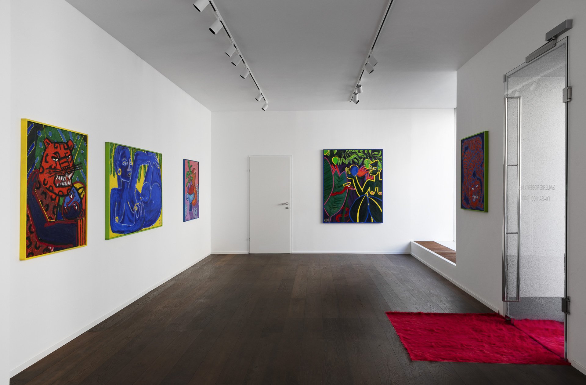 Art gallery with white walls displaying colorful modern paintings, a red carpet near the entrance, and track lighting on the ceiling.
