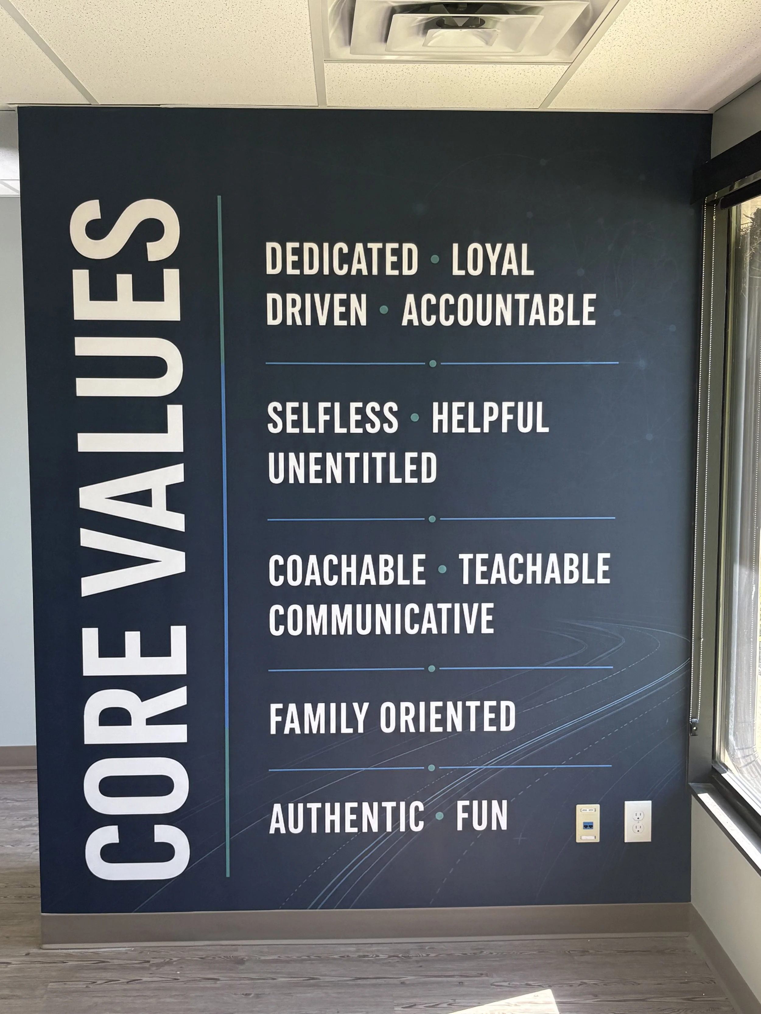 A black wall with the word 'CORE VALUES' written vertically on the left and a list of values including 'DEDICATED • LOYAL', 'DRIVEN • ACCOUNTABLE', 'SELFLESS • HELPFUL', 'UNENTITLED', 'FAMILY ORIENTED', 'AUTHENTIC • FUN', and others.