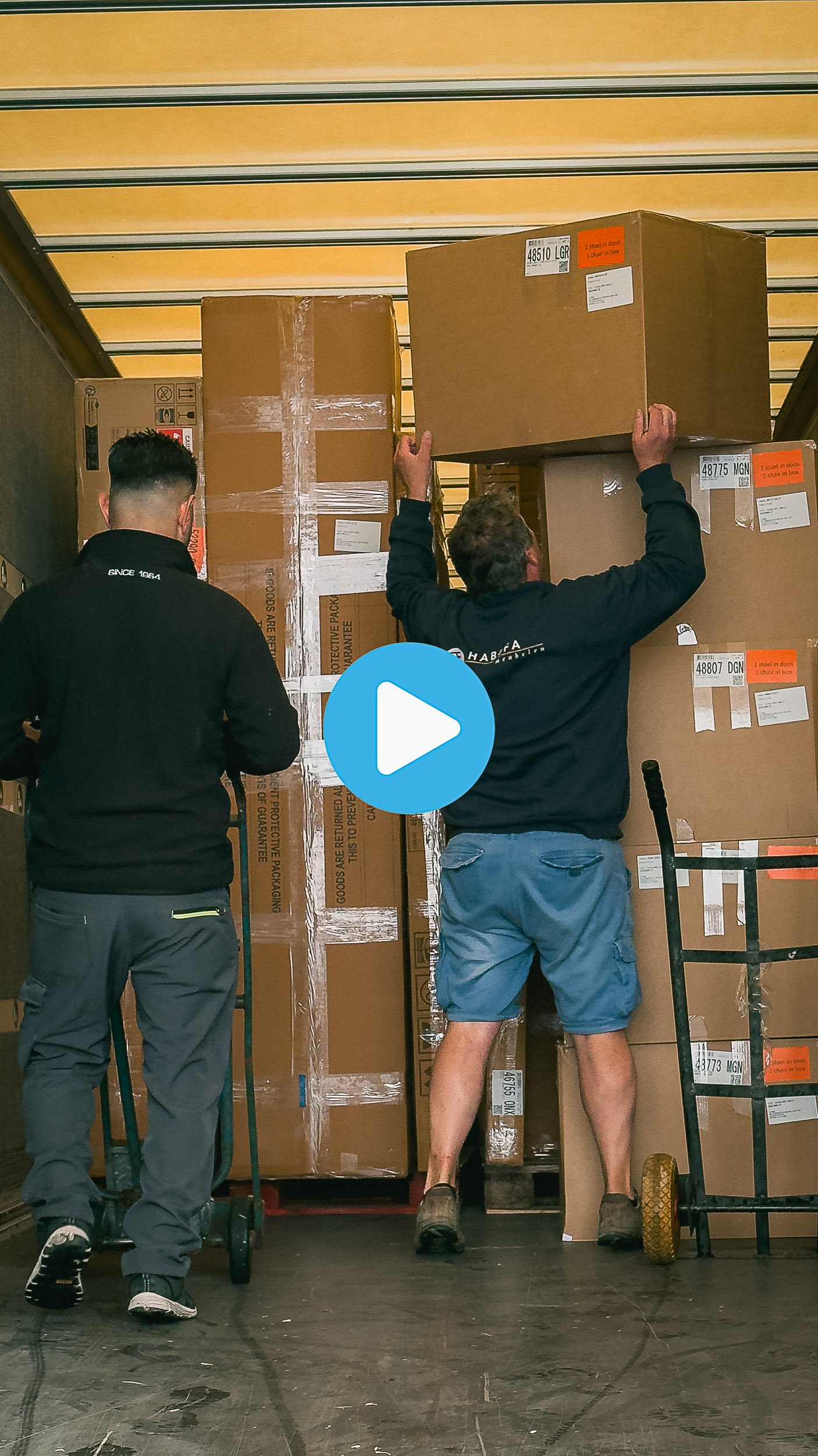 Two workers in a warehouse lift large boxes onto a stack. One worker is wearing a black hoodie, jeans, and sneakers, the other is in a black hoodie, shorts, and work boots. They are surrounded by stacked boxes in a shipping or storage area.