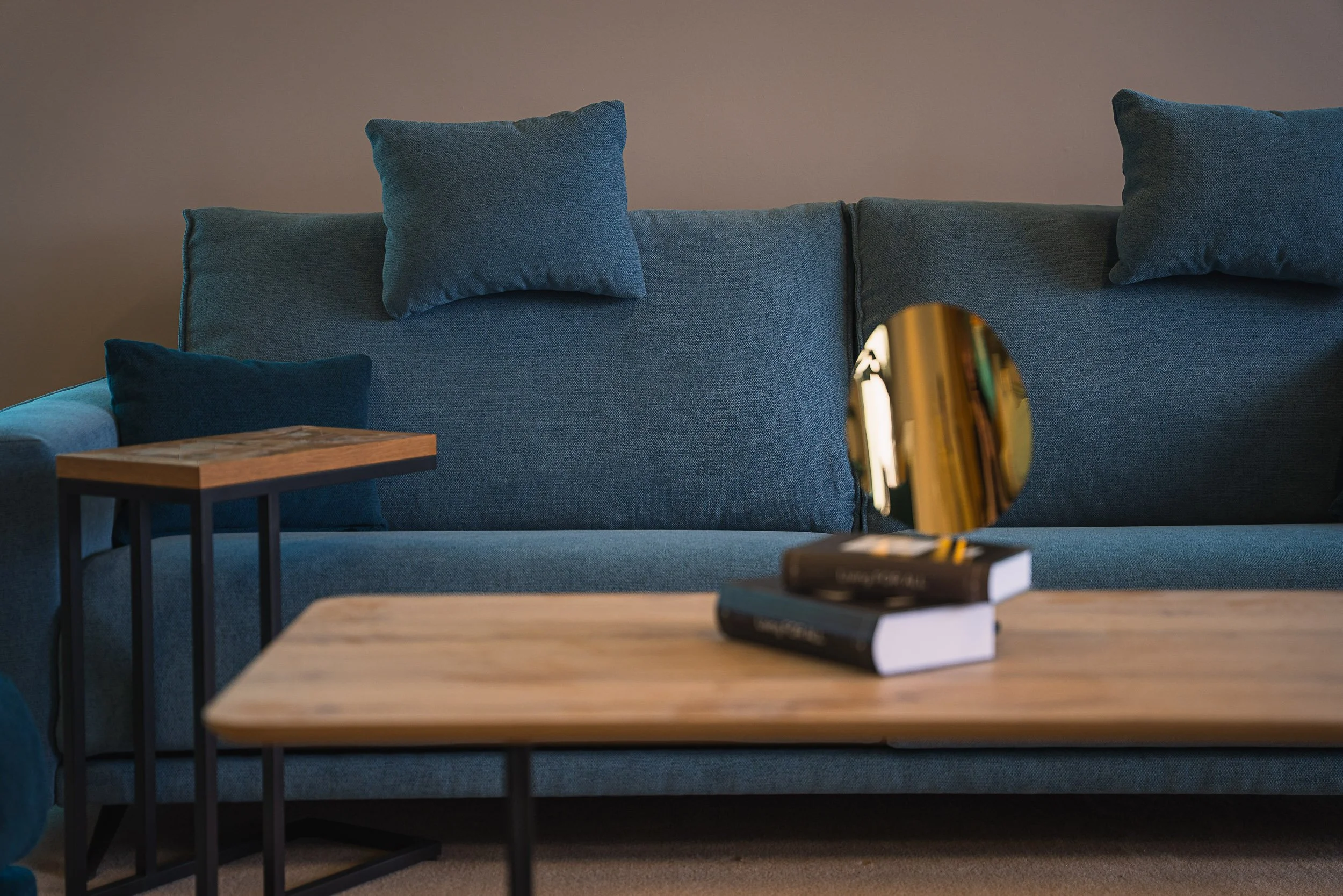 Living room with a blue sofa, matching cushions, a wooden coffee table with two books, a small side table, and a round mirror on top of the books.