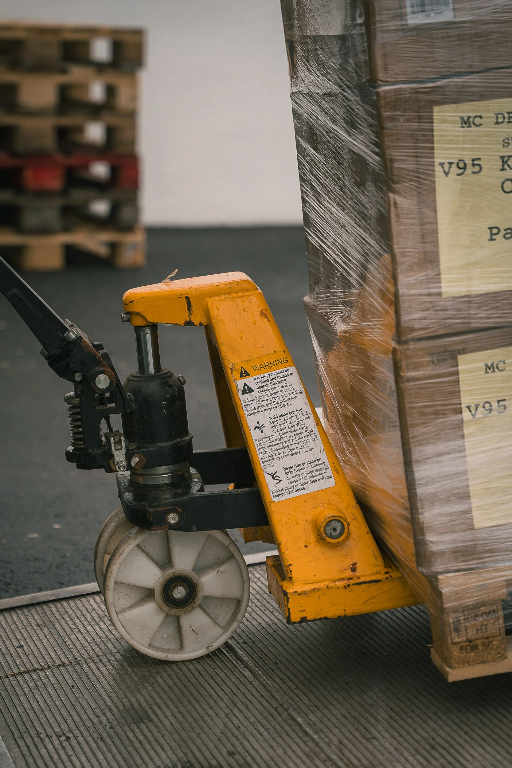 A pallet jack lifting a large pallet of boxes wrapped in plastic, with a stack of pallets in the background.