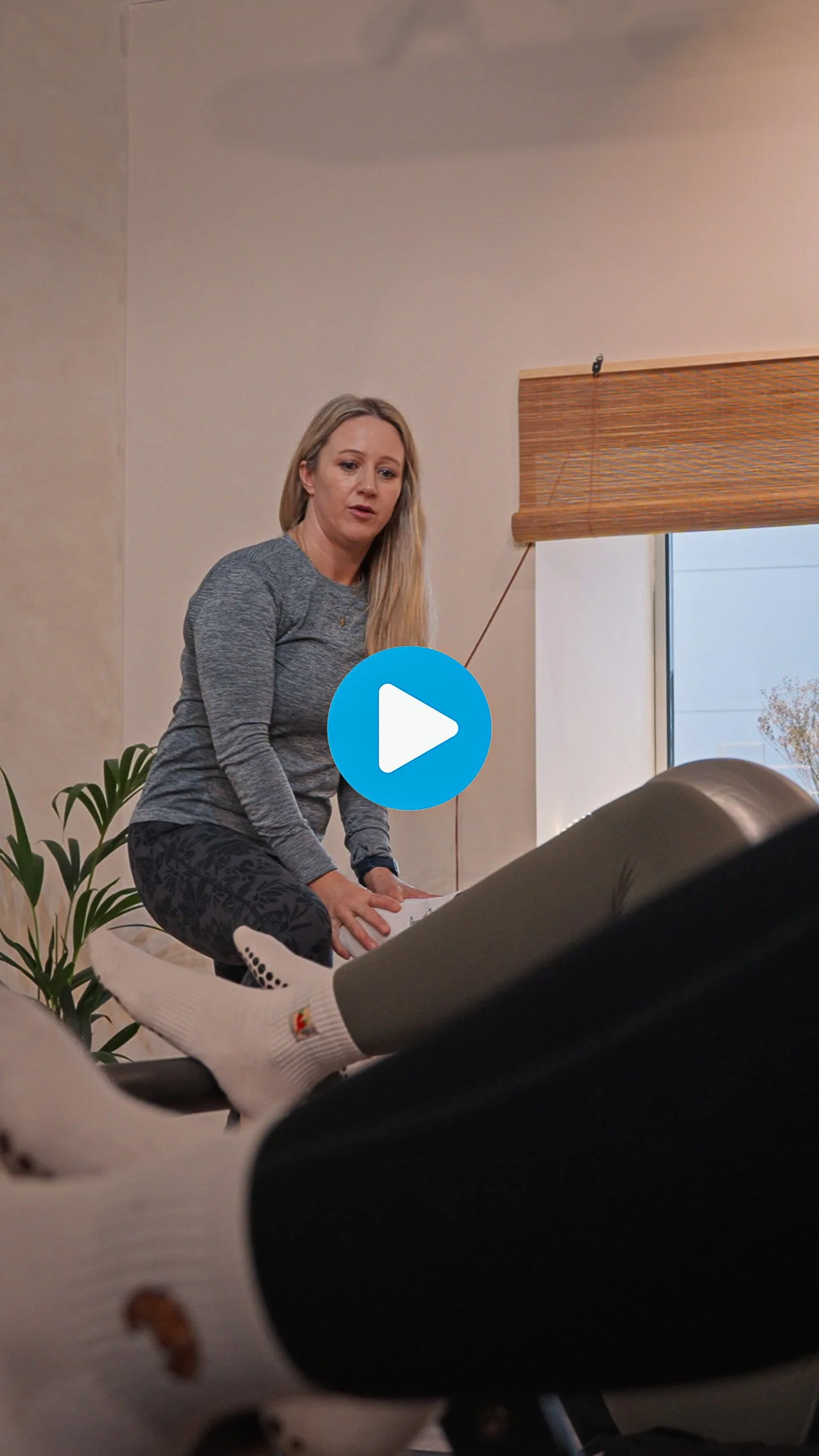 A woman with long blonde hair, wearing a gray long-sleeve shirt and black patterned pants, appears to be talking while sitting on a couch. A person in white socks with a design is sitting with their legs extended in front of her. There is a window wi