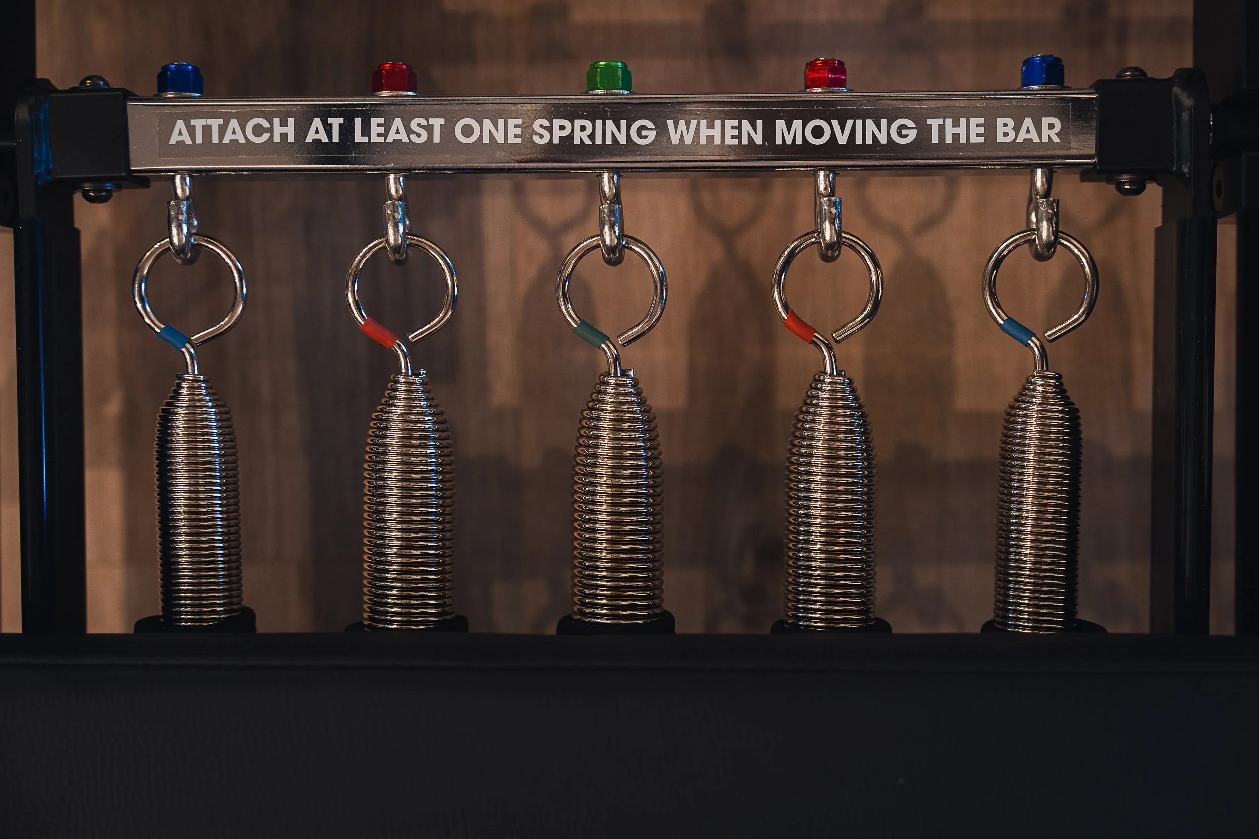A metal gym equipment bar with five coil springs attached to silver rings, each spring connected to a handle. The handles have colored bands at the top: blue, red, green, red, and blue from left to right. There is a black frame supporting the bar, wi