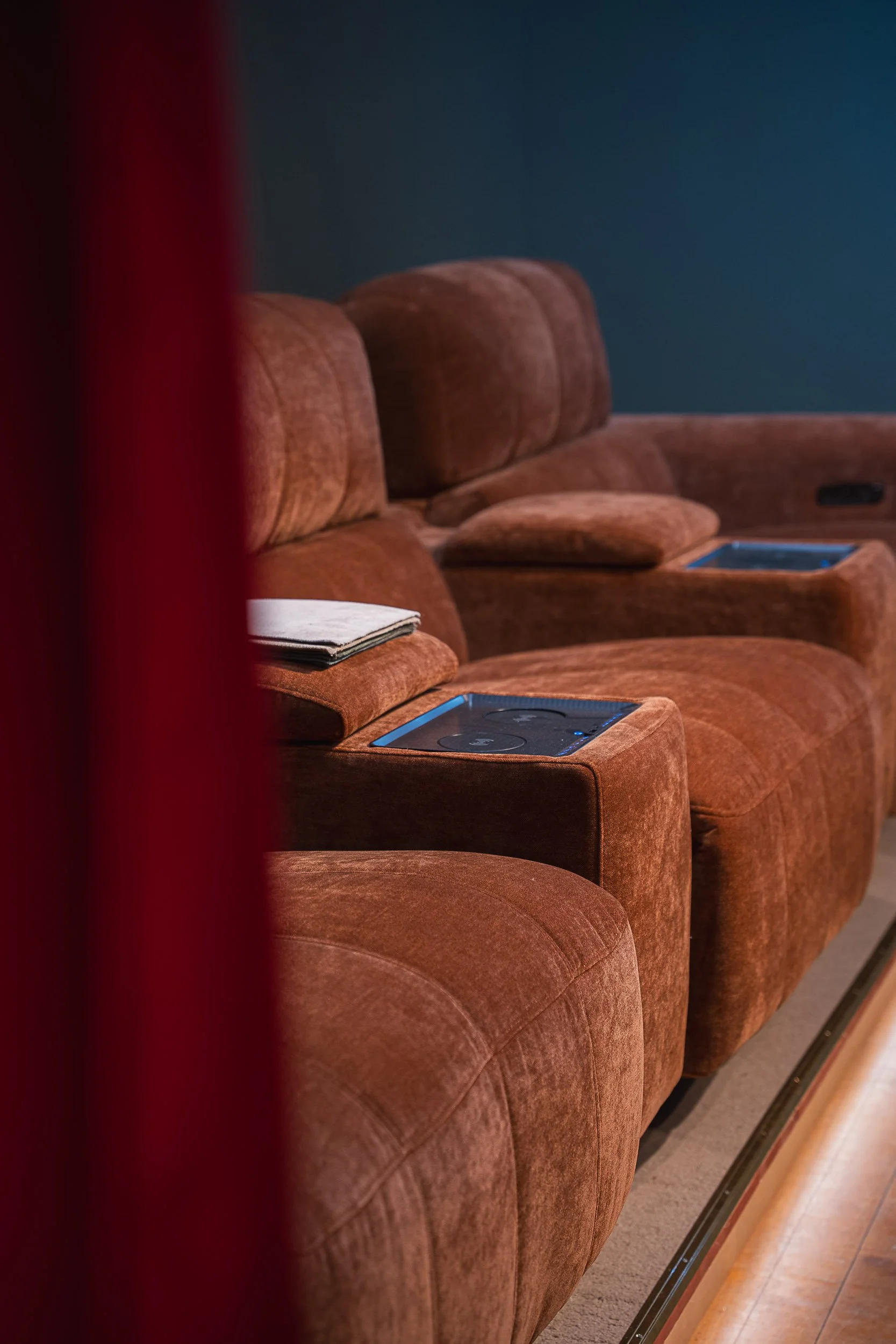 A row of brown plush theater seats with built-in tablet screens on the armrests, seen from a slightly side angle, with a blue wall in the background.