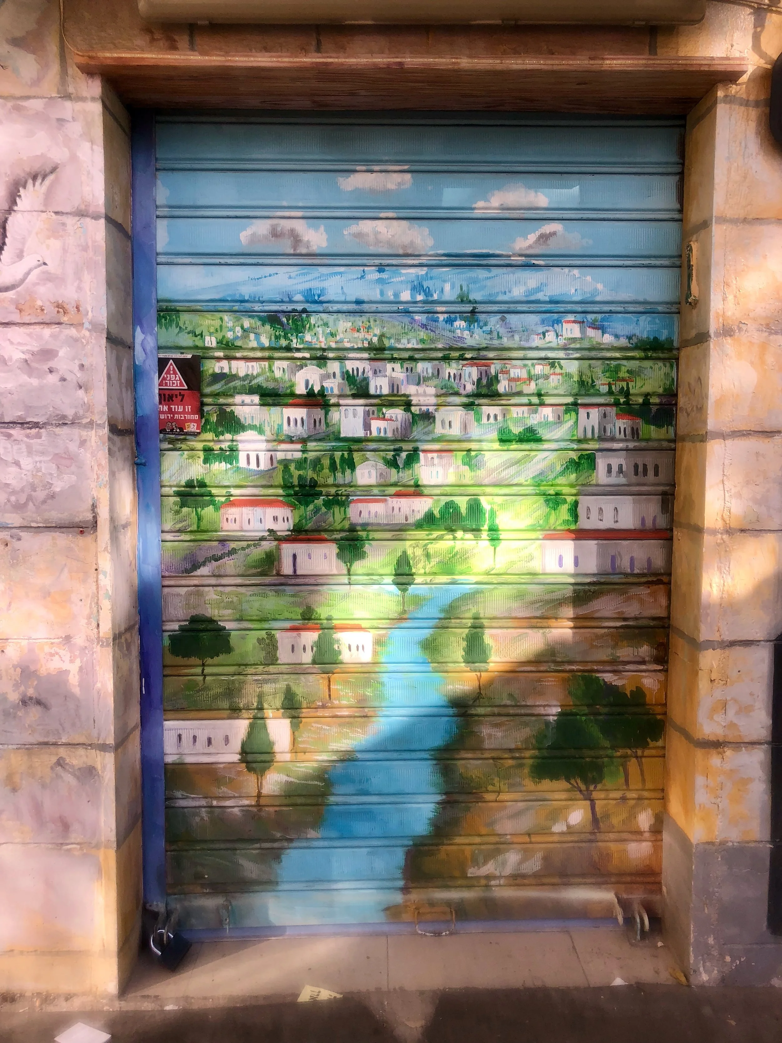 Mural painted on a metal roll-up door depicting a serene landscape with a stream flowing through green hills, trees, and a village with white buildings and red roofs under a cloudy sky.