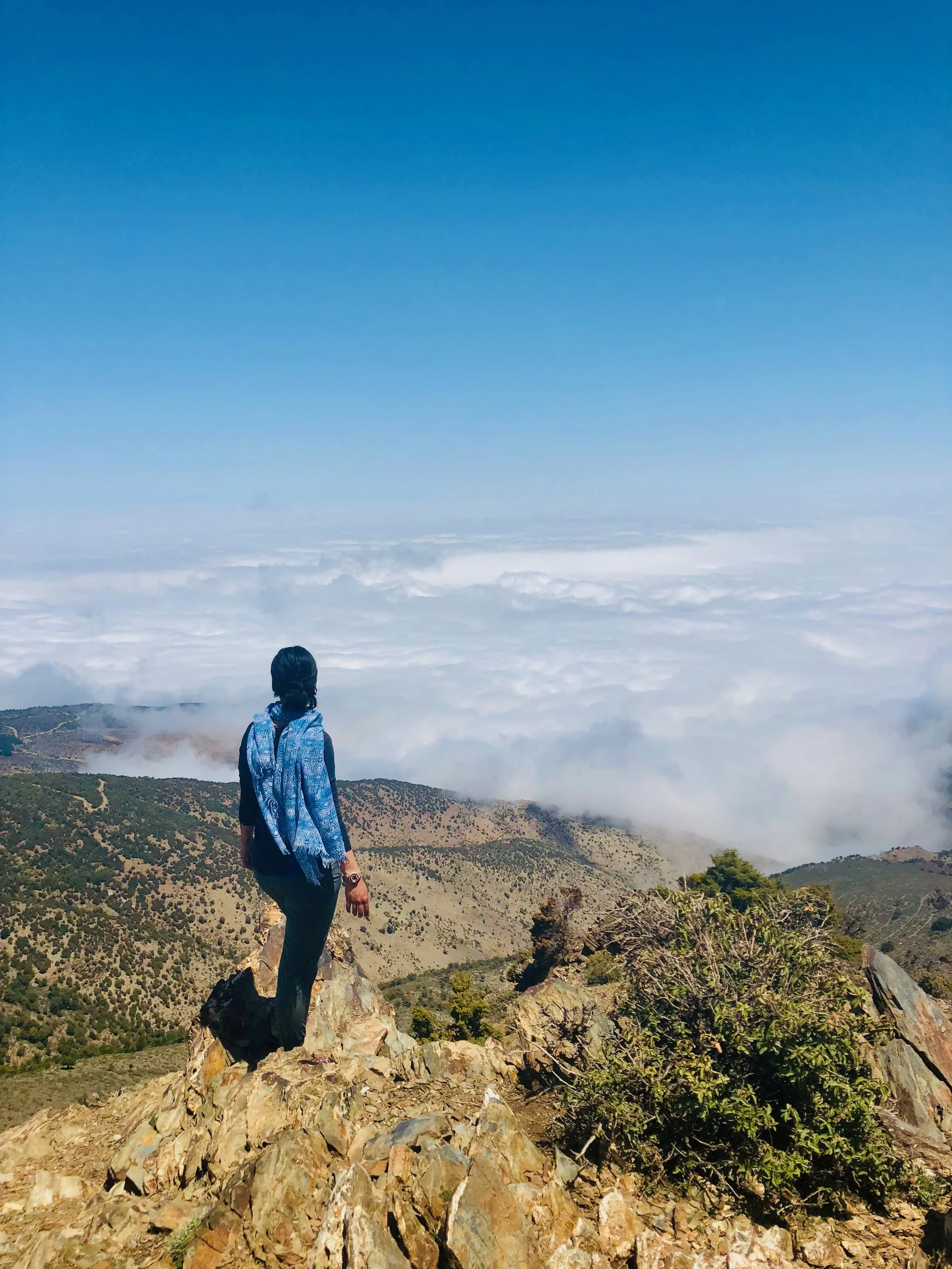 Person standing on rocky mountain overlooking a cloud-filled valley under a clear blue sky.