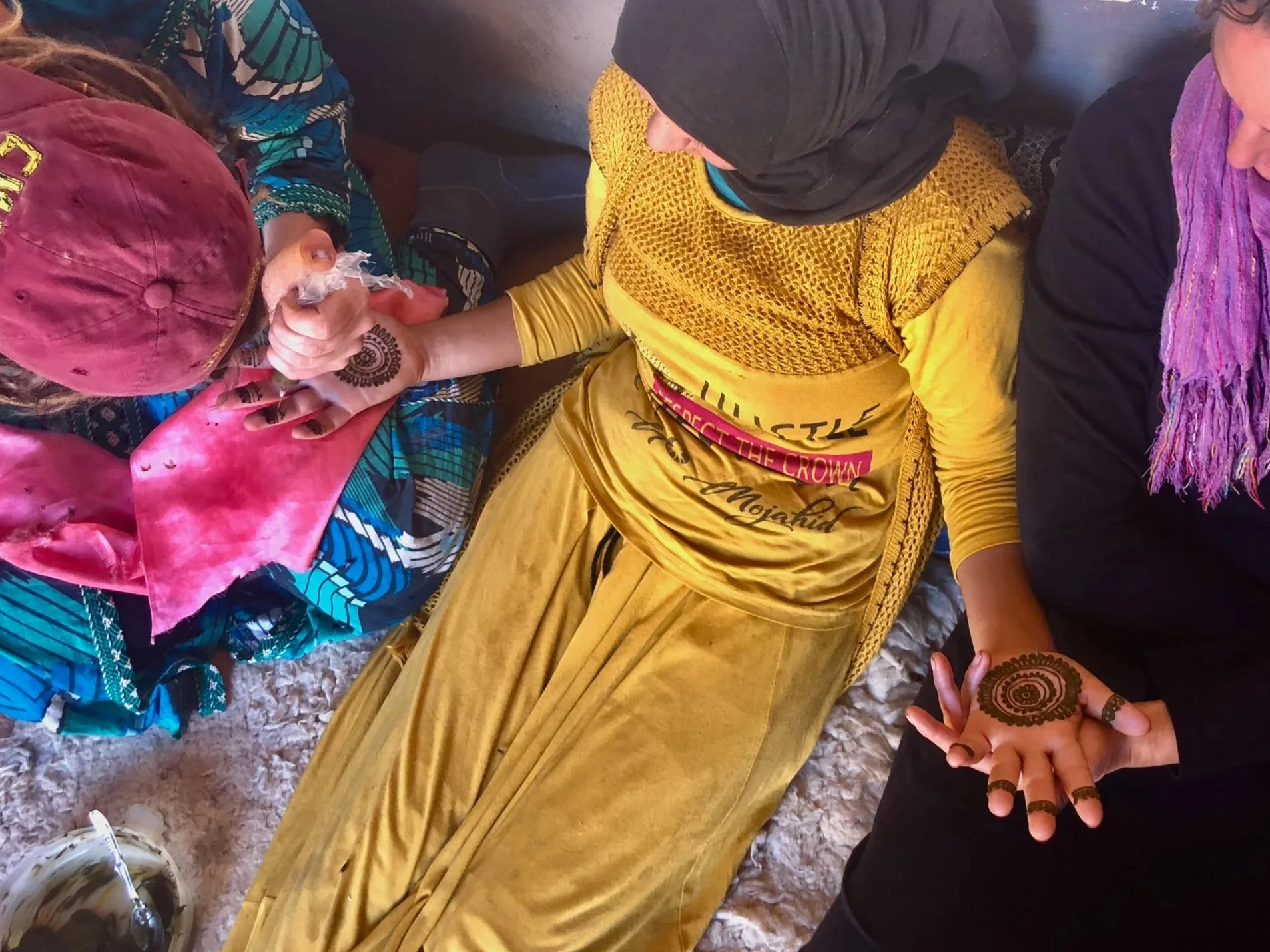 Three women sitting on a plush carpet, two showing off henna designs on their hands.
