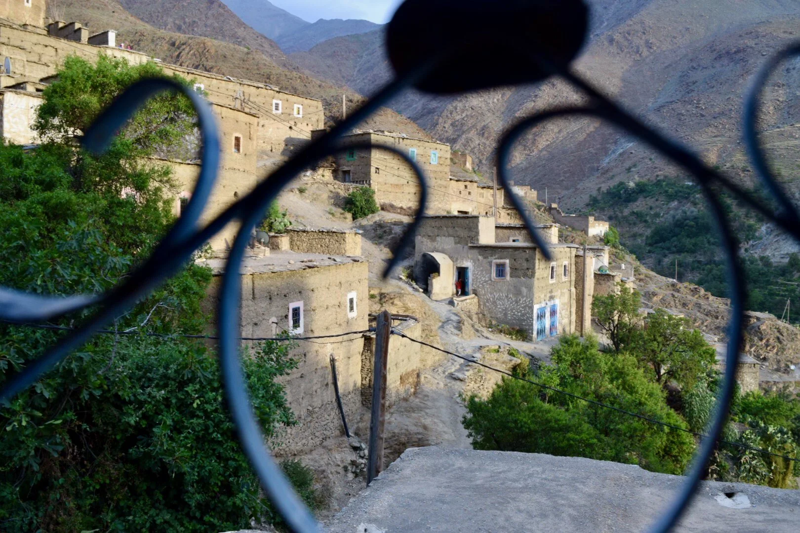 Mountain village with adobe-style houses on a hillside, seen through a decorative iron fence in the foreground, with mountains in the background.