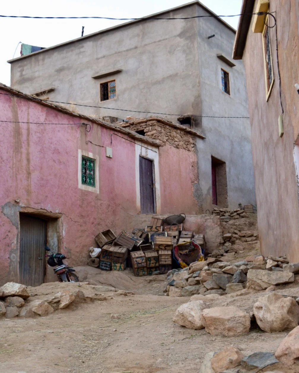 A rustic street scene showing old buildings with pink and gray walls, some with small windows. There are scattered rocks and a pile of wooden crates on the uneven, dirt ground. A motorcycle is parked near a doorway on the left. Overhead electrical wires are visible against the sky.