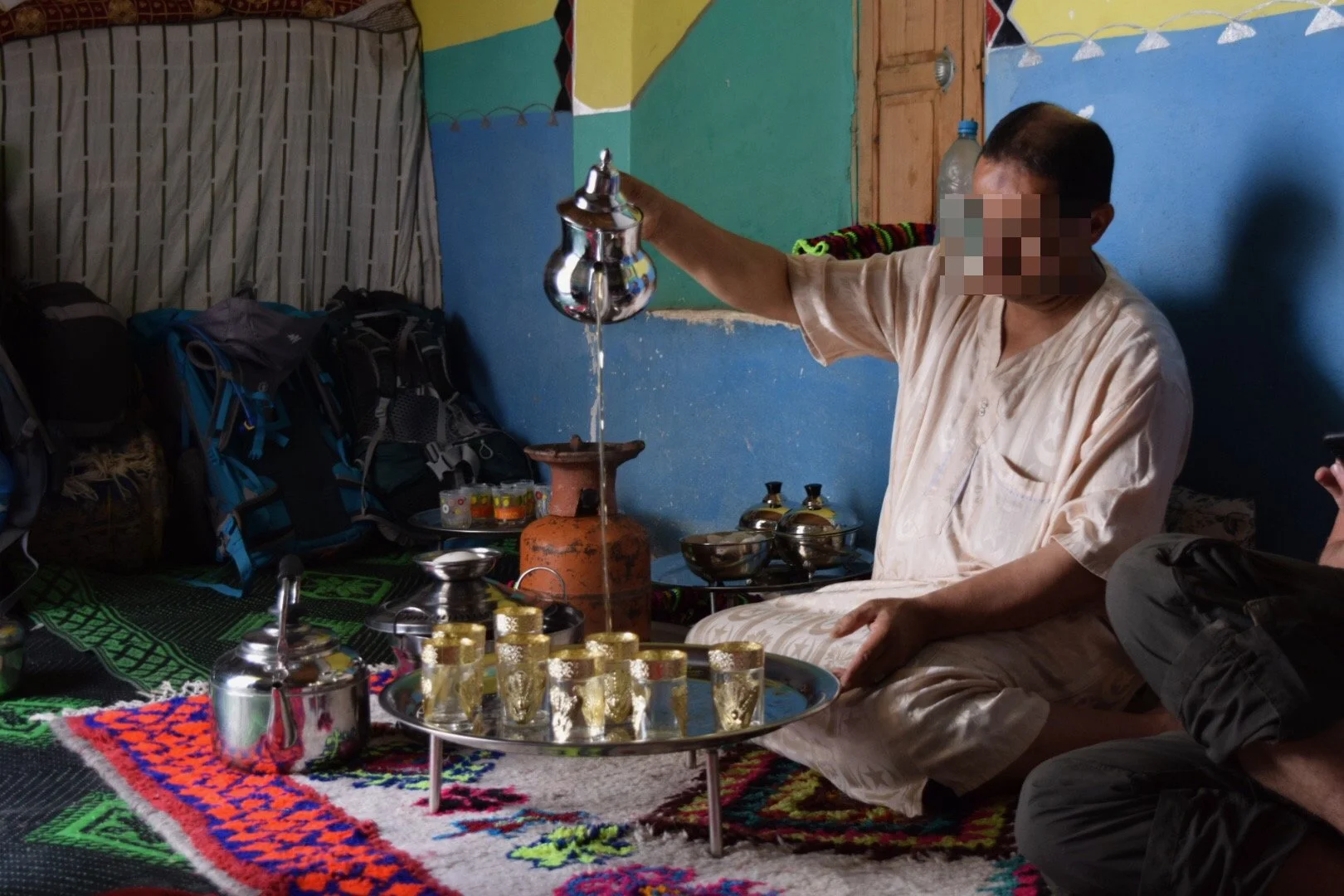Man in traditional attire performing a ritual with a metal vessel pouring water into a clay lamp while others observe, seated on a colorful carpet inside a decorated room.