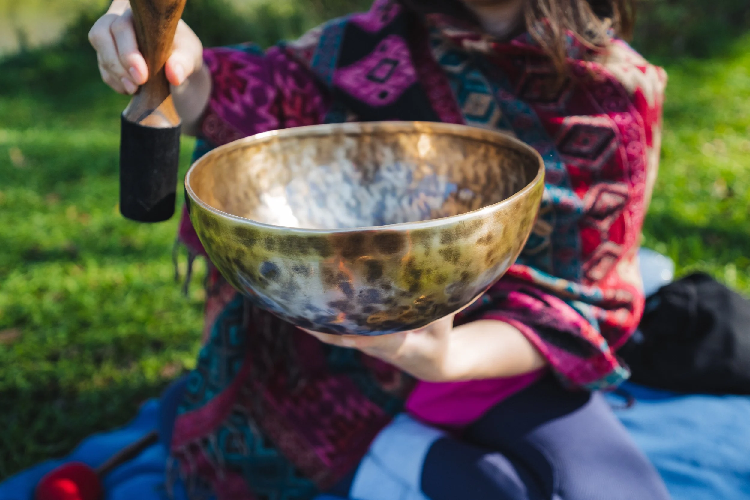 Person sitting outdoors playing a singing bowl with a mallet in a grassy area.