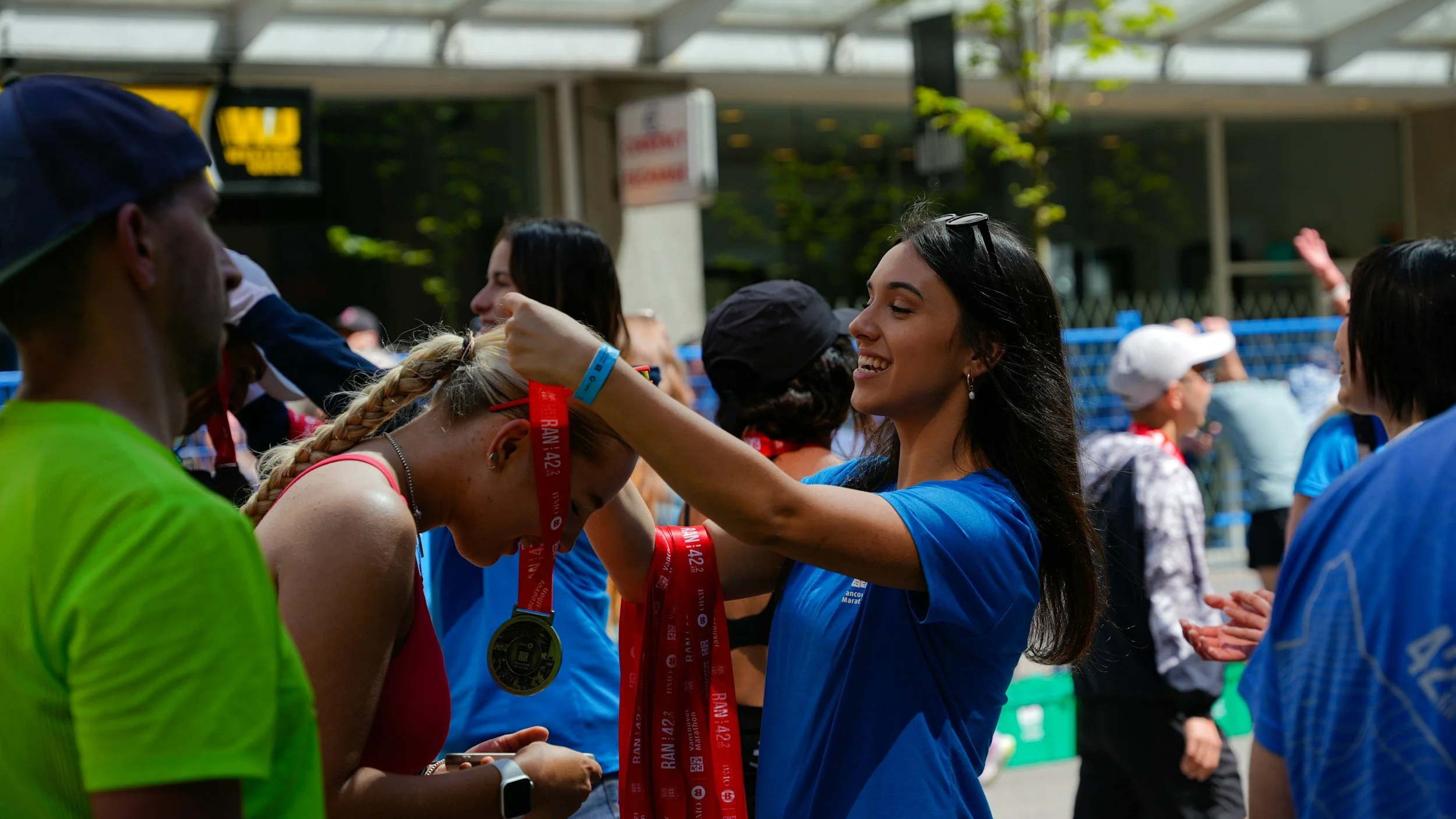 A woman in a blue shirt medals a female marathon runner with blonde hair in a braid, who is wearing a pink top. The runner is bowing her head as she receives the medal, surrounded by other runners and spectators at an outdoor event.