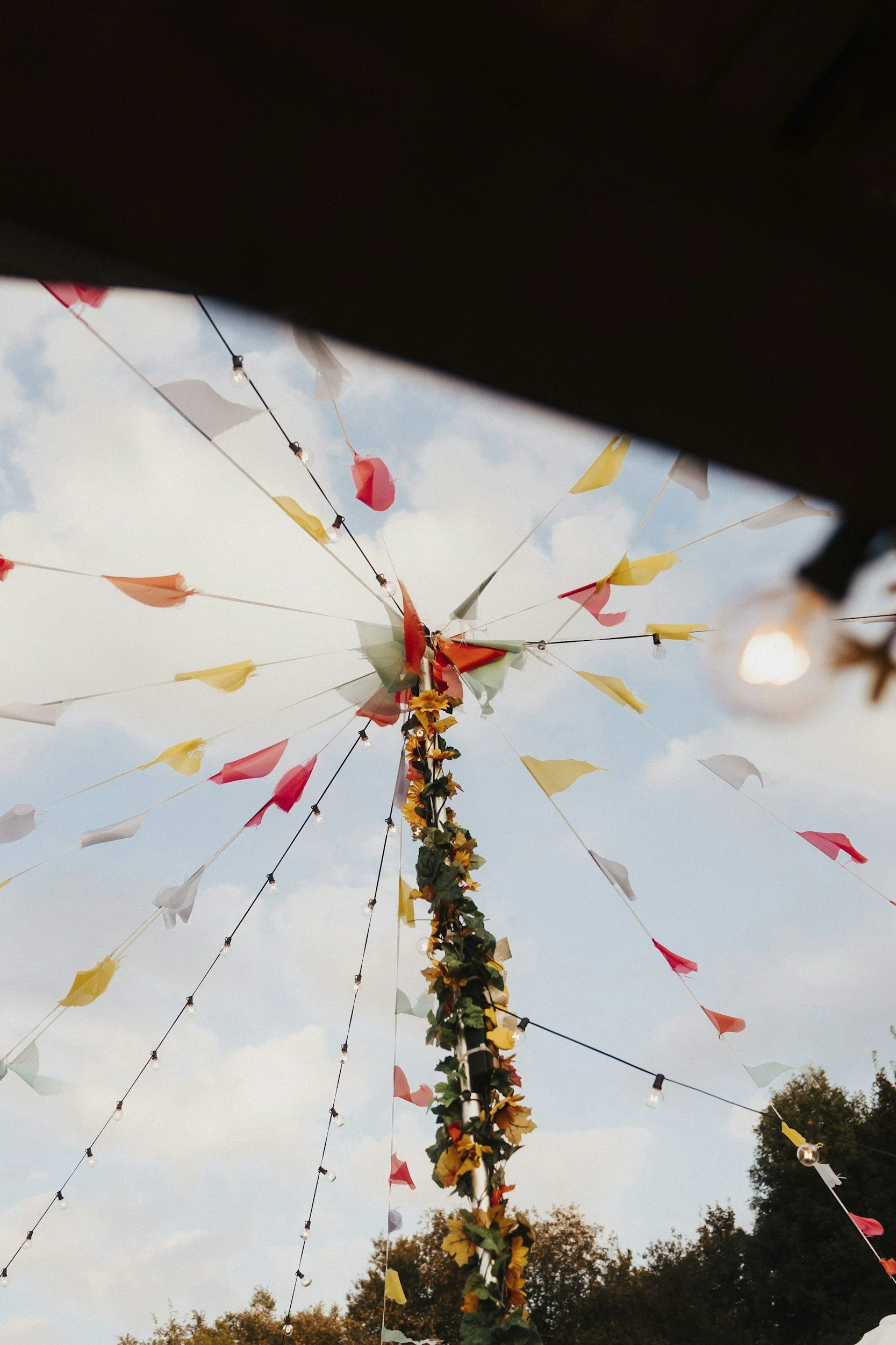 Colorful outdoor decoration with flags and string lights on a pole, under a partly cloudy sky, with trees in the background.