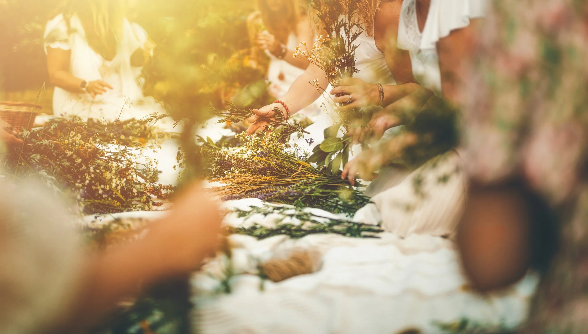 People gathered outdoors arranging and picking wildflowers on a long cloth-covered table, dressed in white, during daylight.