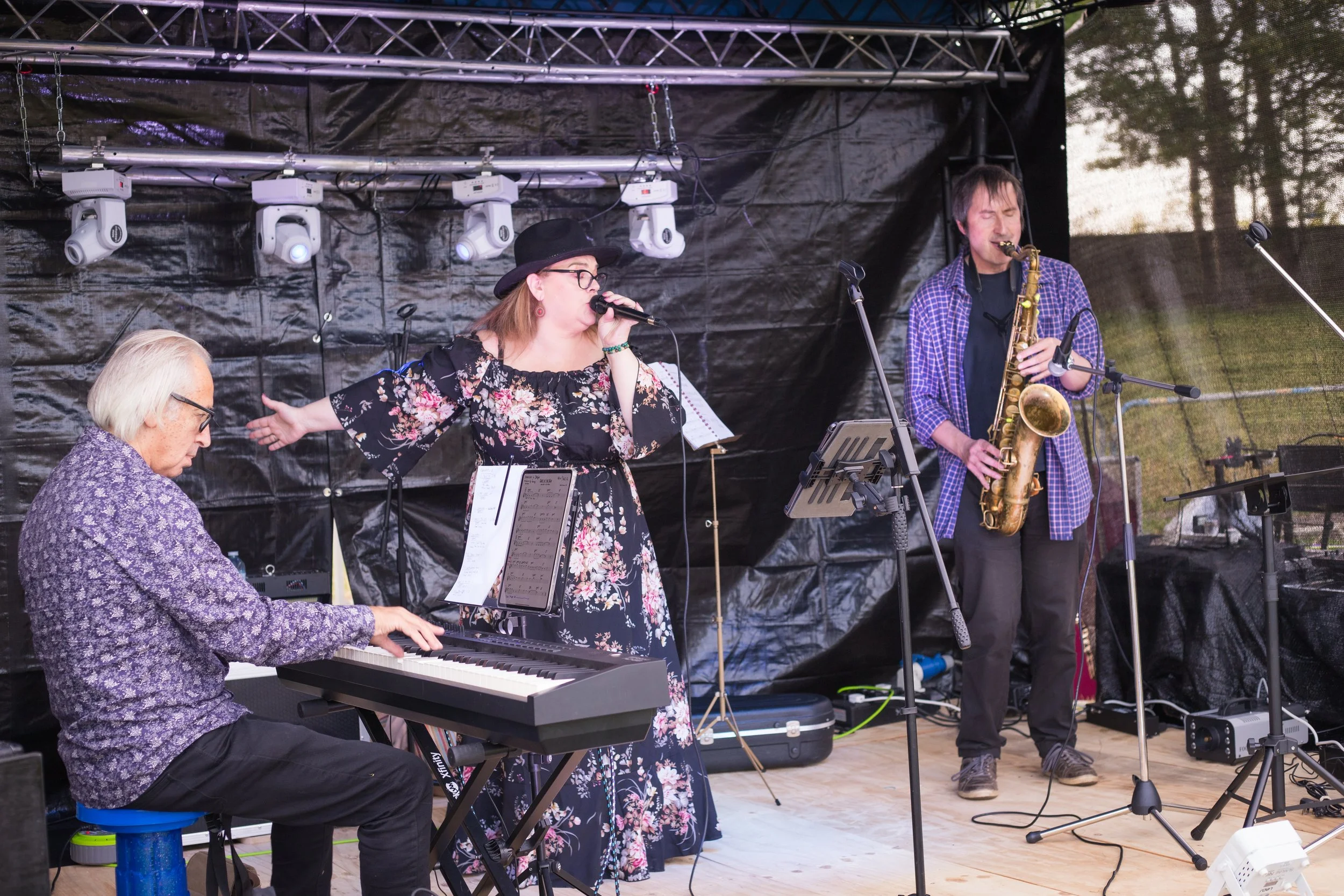 Three musicians performing on stage outdoors: a pianist, a female vocalist, and a saxophonist. The stage has black backdrop and lighting equipment overhead.