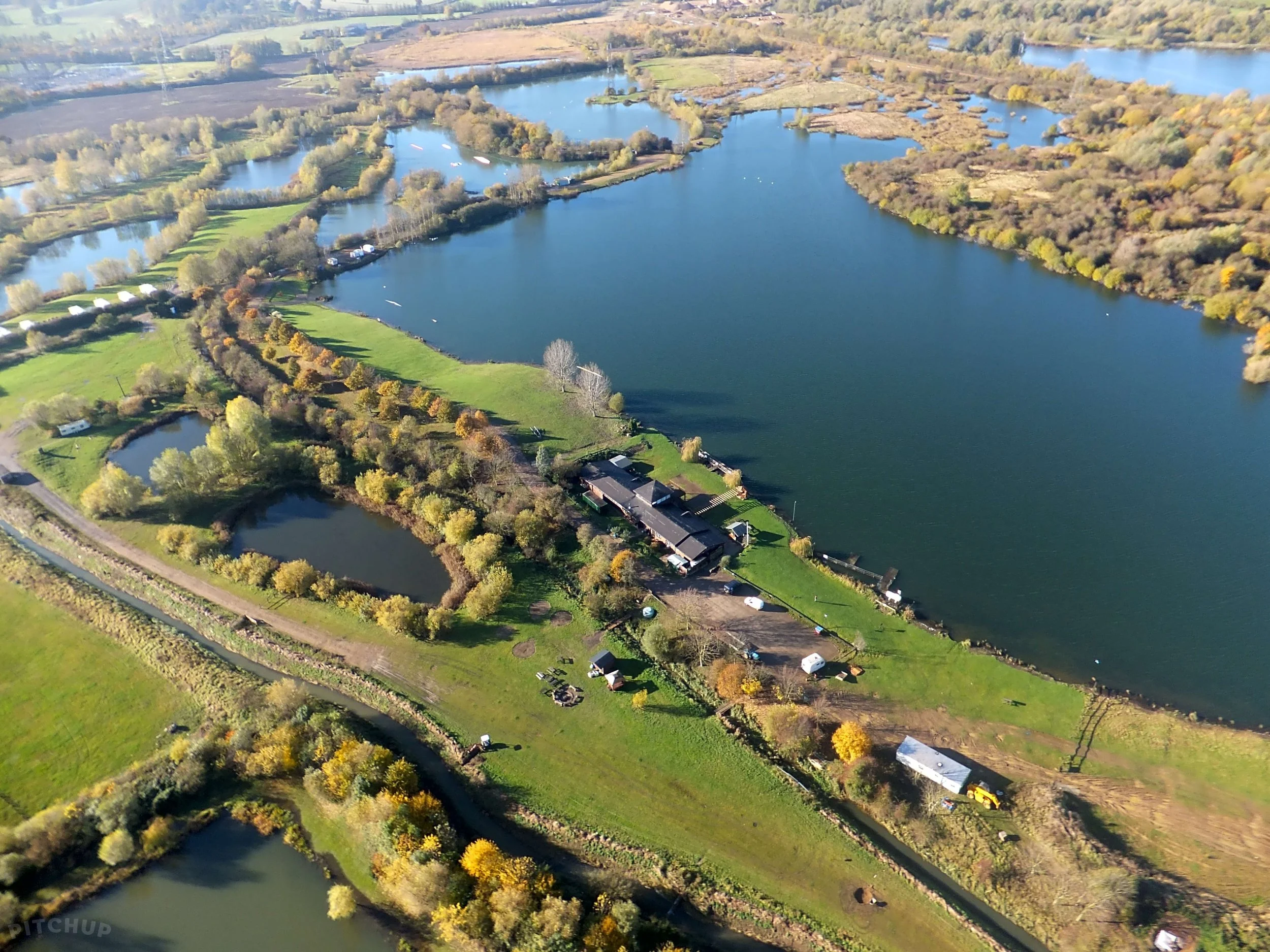 Aerial view of a large lake with surrounding greenery, trees, and some buildings and boats on the water.