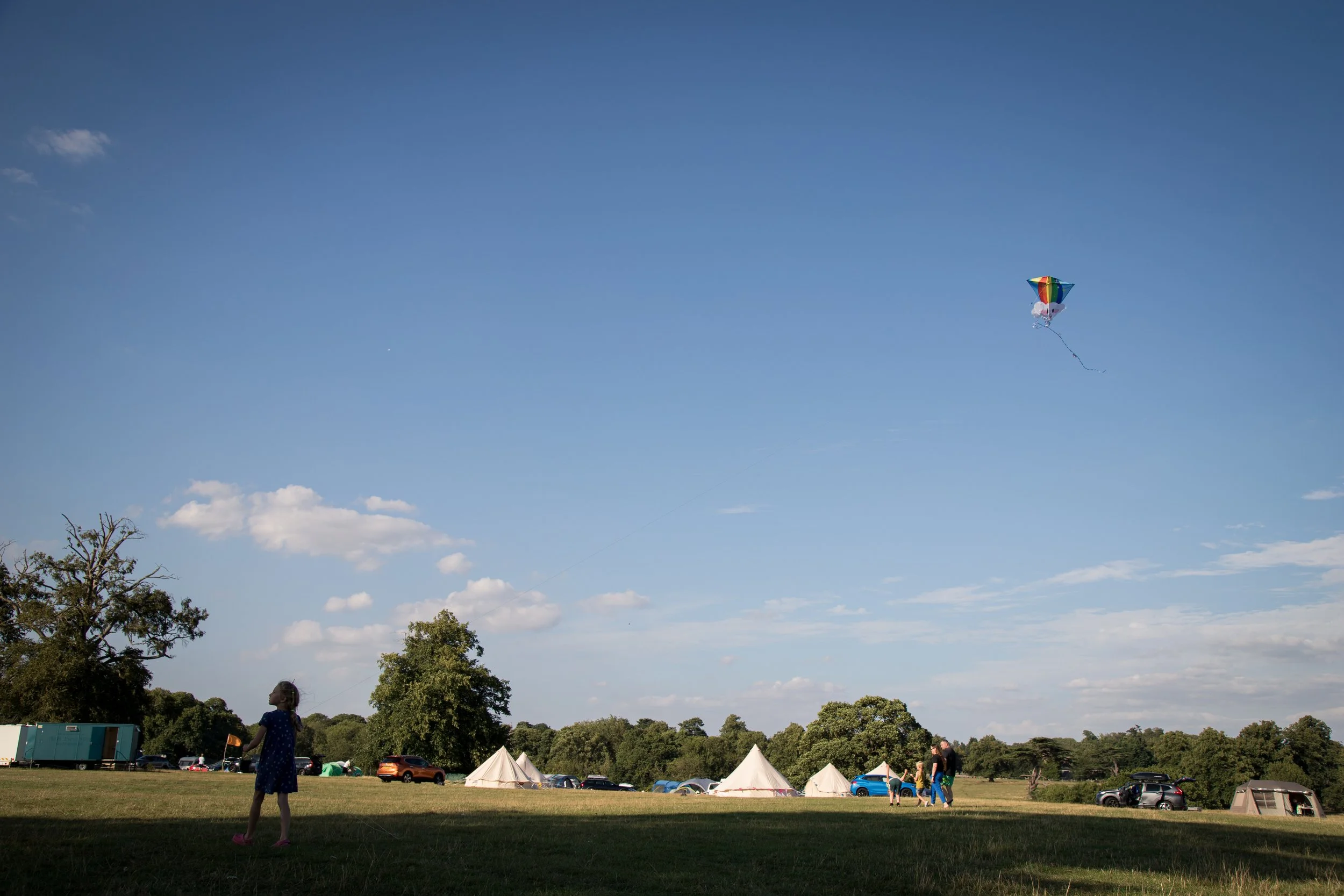 A girl flying a colorful kite in a grassy field during daytime, with tents and cars in the background under a partly cloudy sky.