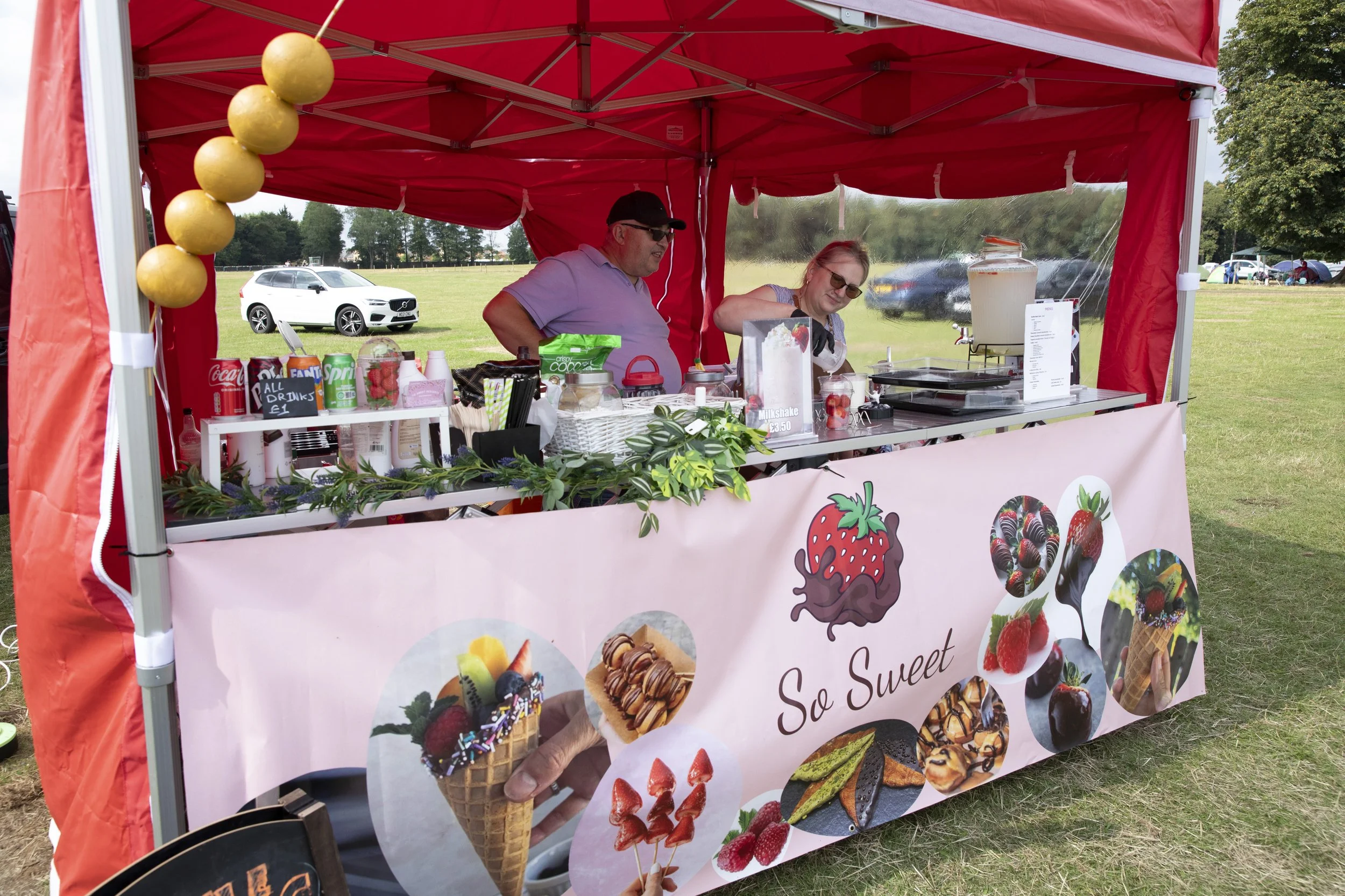 A vibrant outdoor stall with a pink banner reading 'So Sweet' displaying various ice cream and dessert images. Two people are inside the stall, a man and a woman, preparing treats. The stall is under a red canopy with a string of yellow decor. The ba
