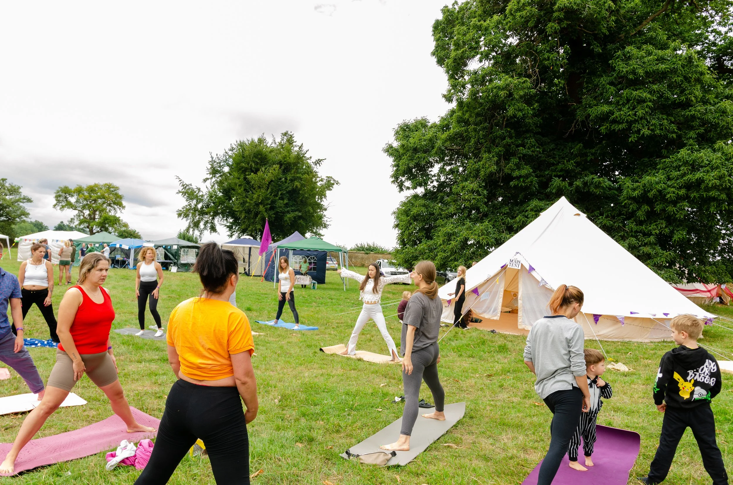 Group of people participating in an outdoor yoga class on a grassy field, with tents and trees in the background.