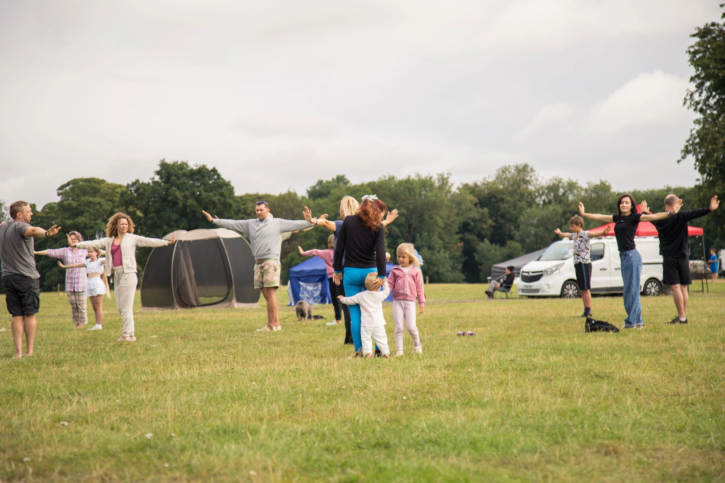 People standing in a circle with arms outstretched on a grassy field during an outdoor gathering or festival, with tents, a van, and trees in the background.
