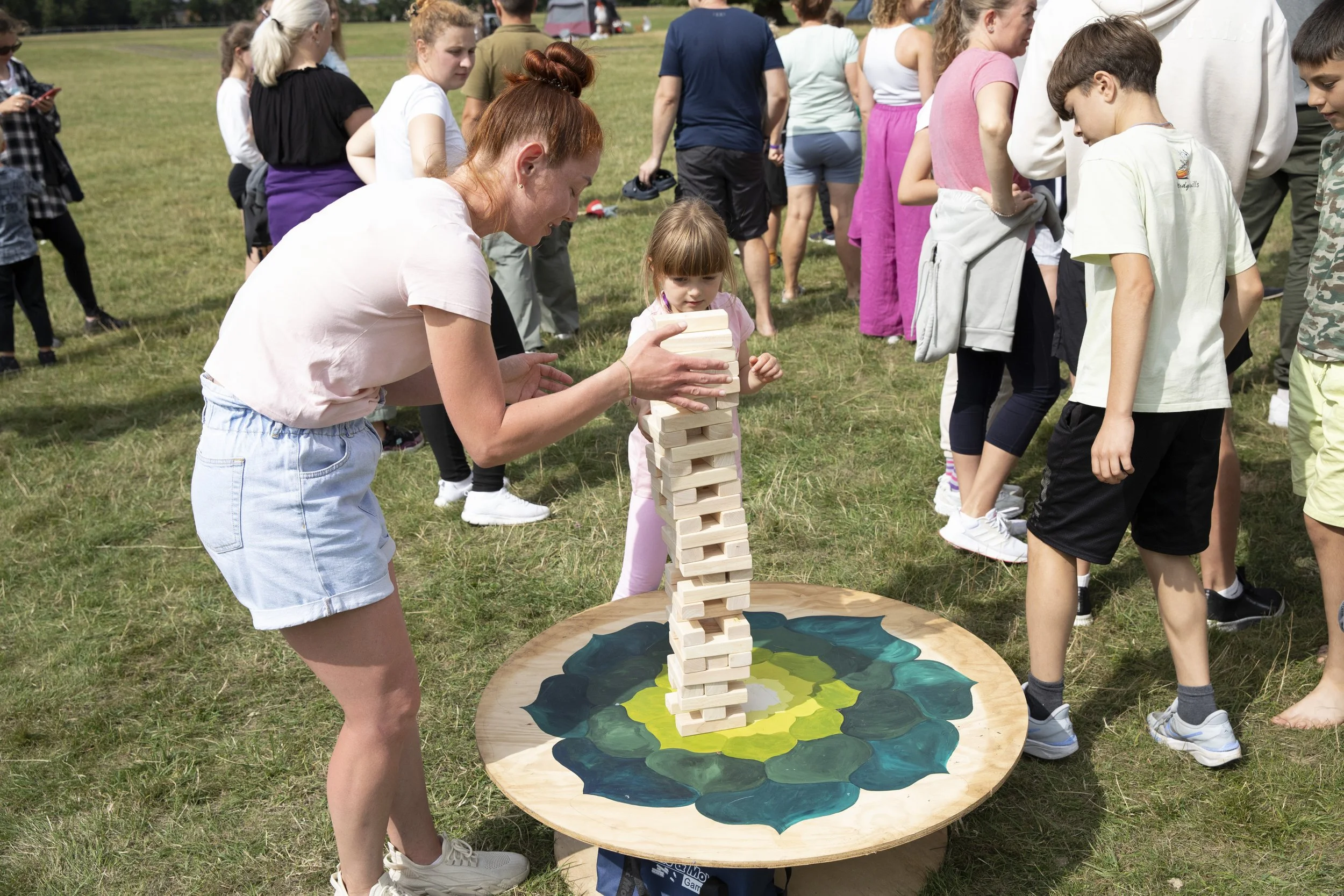 A woman and a young girl are playing Jenga outdoors on a grassy field, with other people standing in line or watching in the background.