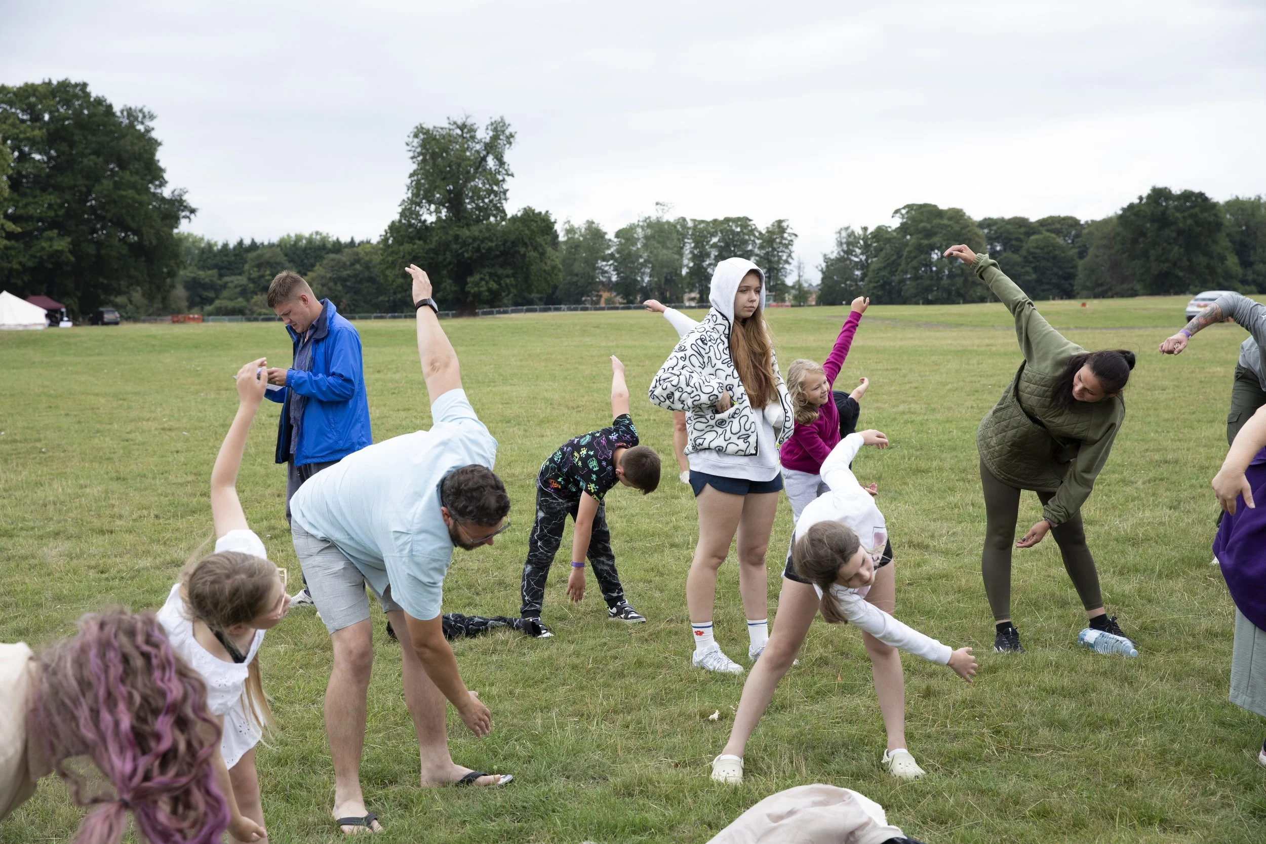 A group of children and adults stretching outdoors on a grassy field with trees in the background, some wearing casual clothing and hoodies.