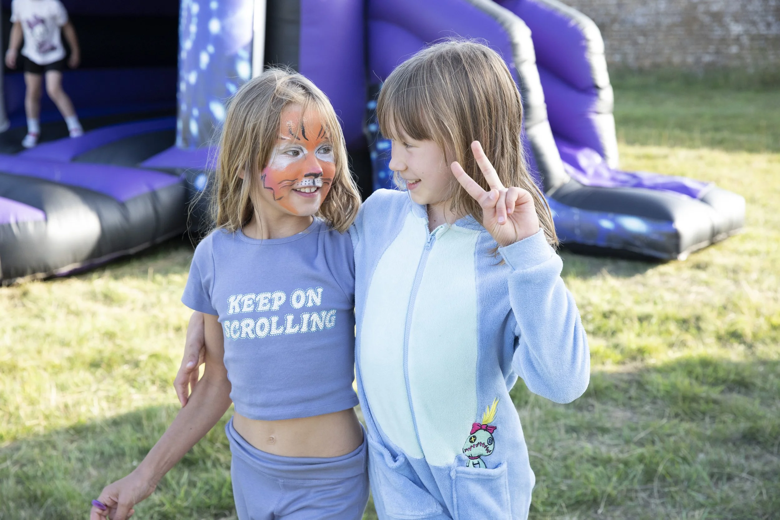 Two young girls at an outdoor bounce house, one with face paint resembling a tiger and making a peace sign, smiling at each other.