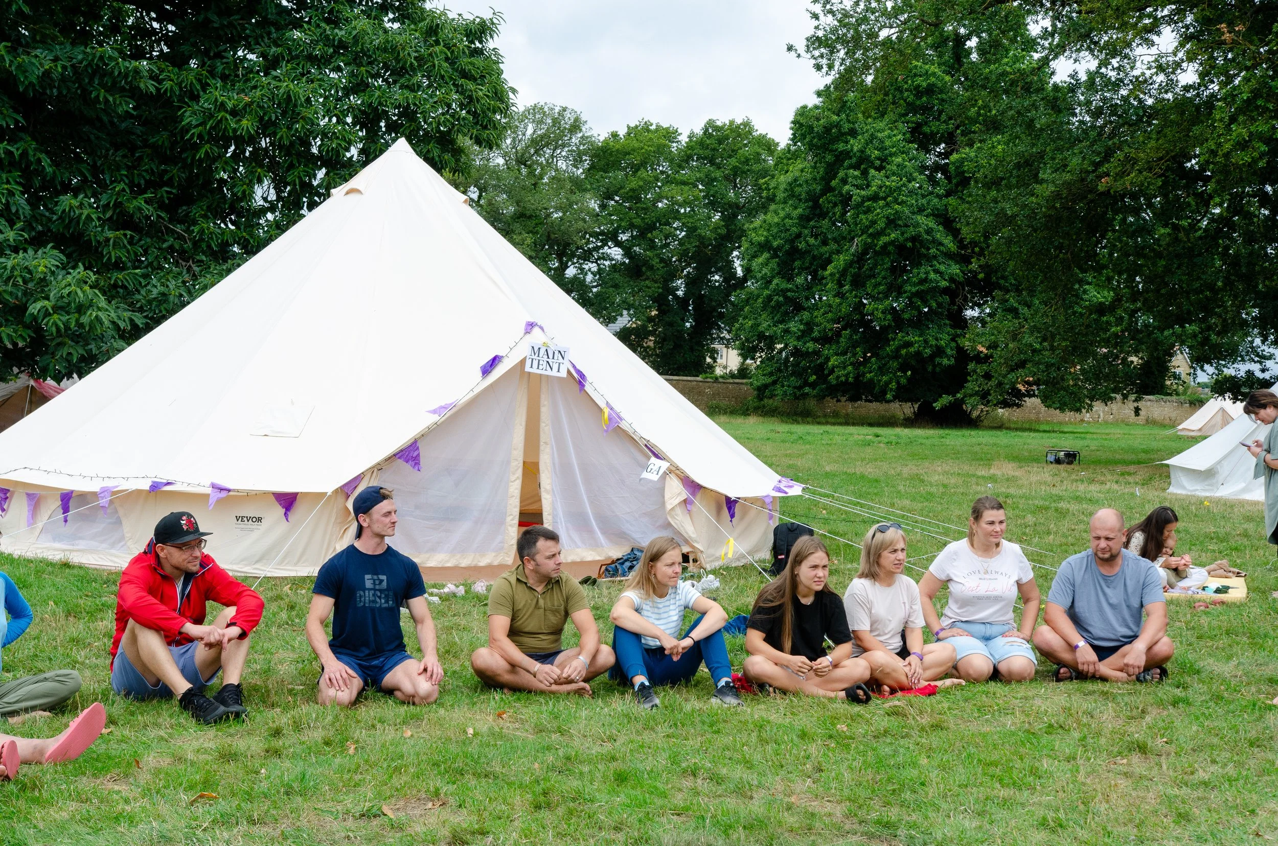 A group of nine people sitting on the grass in front of a large white tent outdoors, with trees in the background.