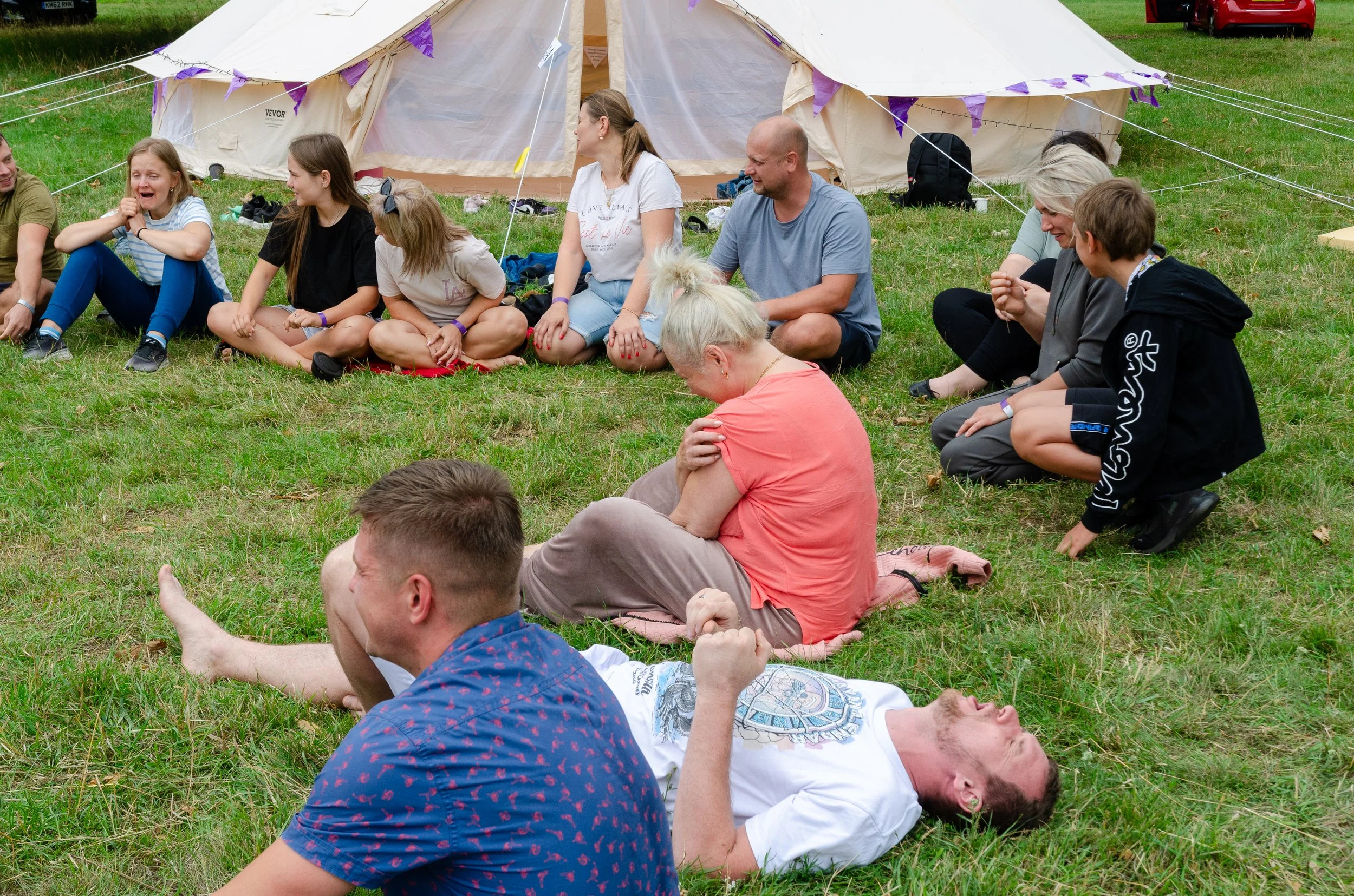 Group of people sitting and lying on grass in front of a large tent, engaging in conversation and laughter during an outdoor event.