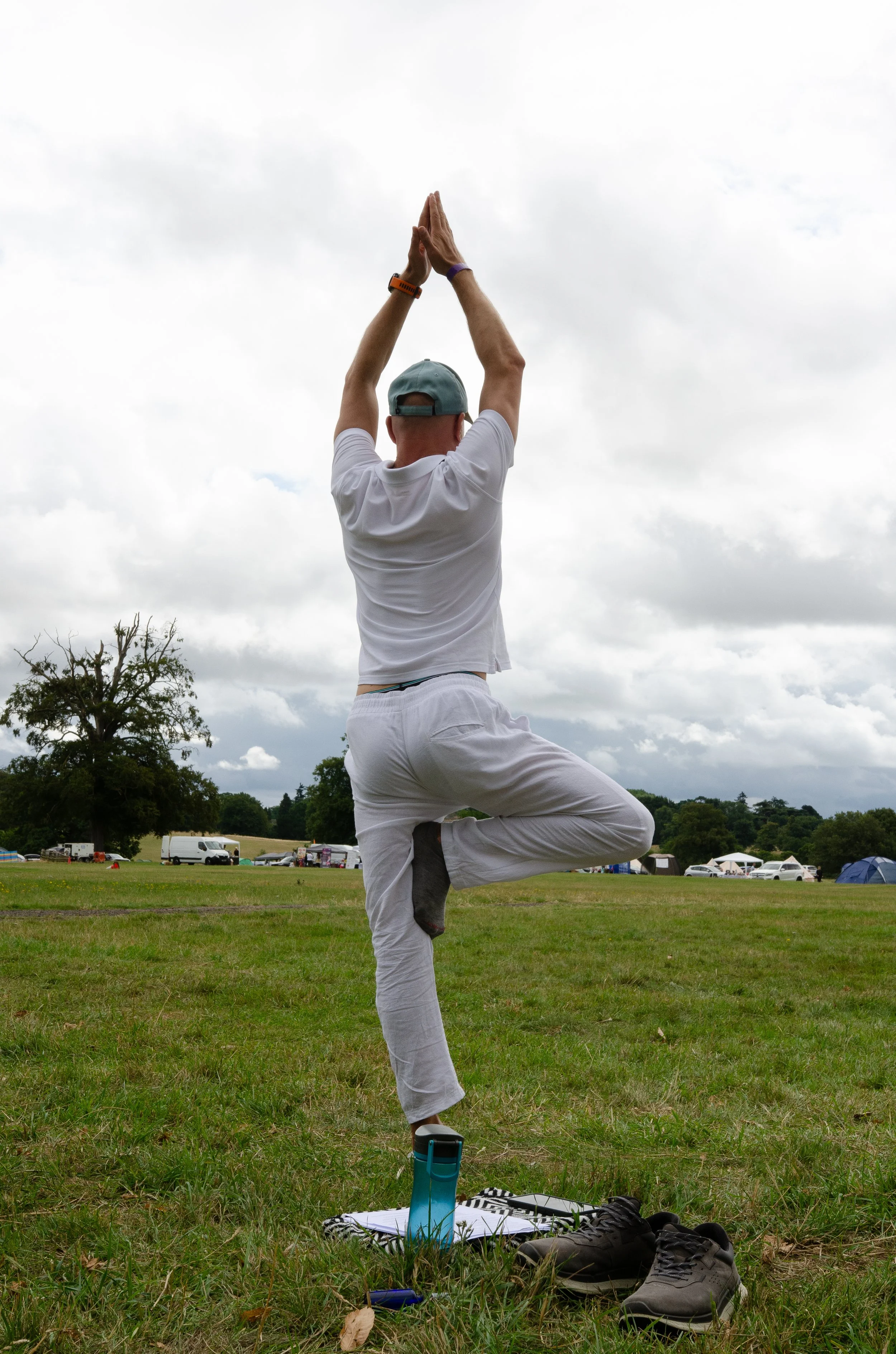 A person practicing yoga outdoors on a cloudy day, standing on one leg in a tree pose with hands in prayer position above their head, on a grassy field with tents and vehicles in the background.