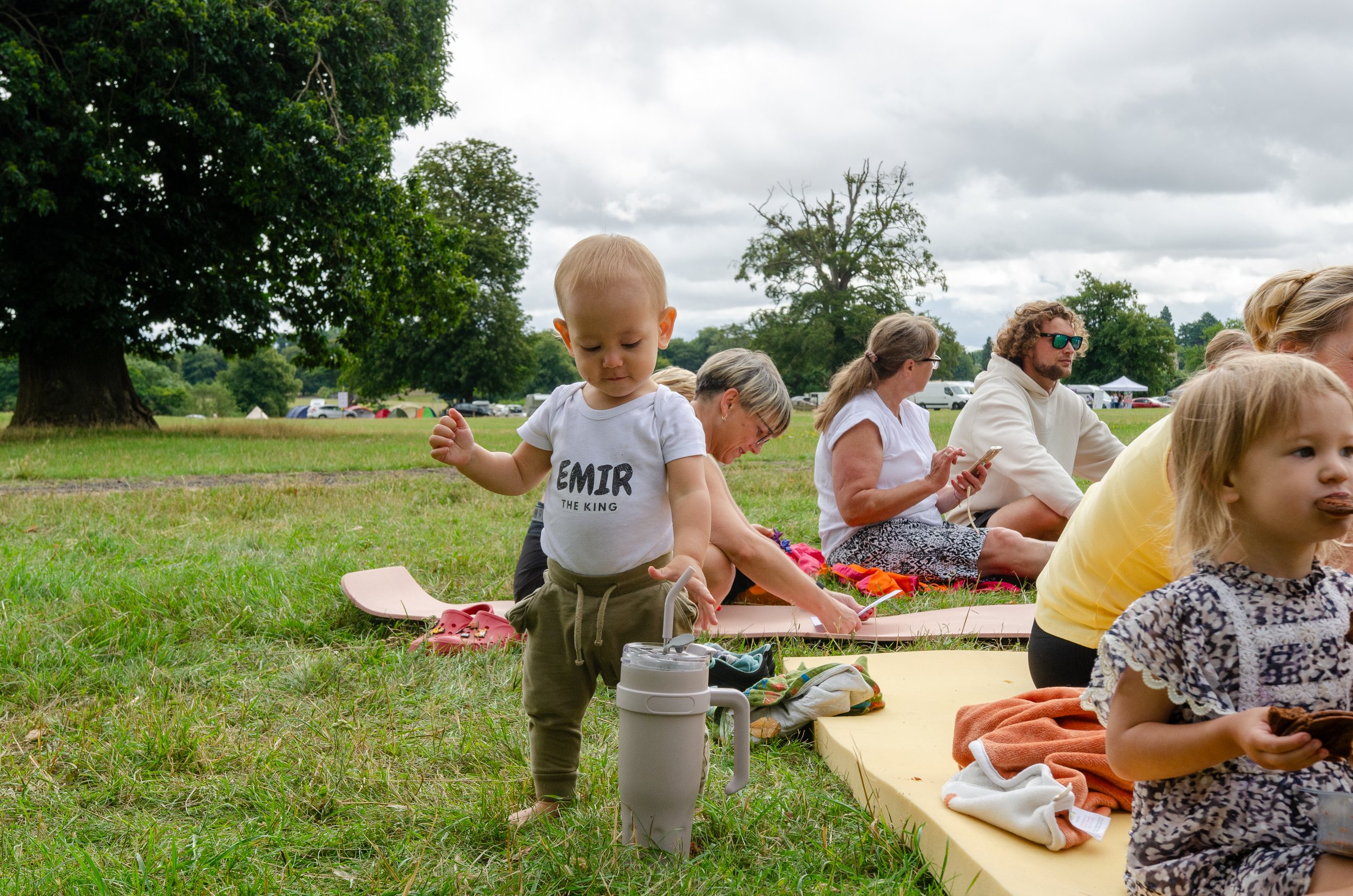 Children and adults sitting on mats and blankets in a grassy park, with trees and tents in the background on a cloudy day. A young boy in a white T-shirt with the words 'EMIR THE KING' is standing near a water bottle.