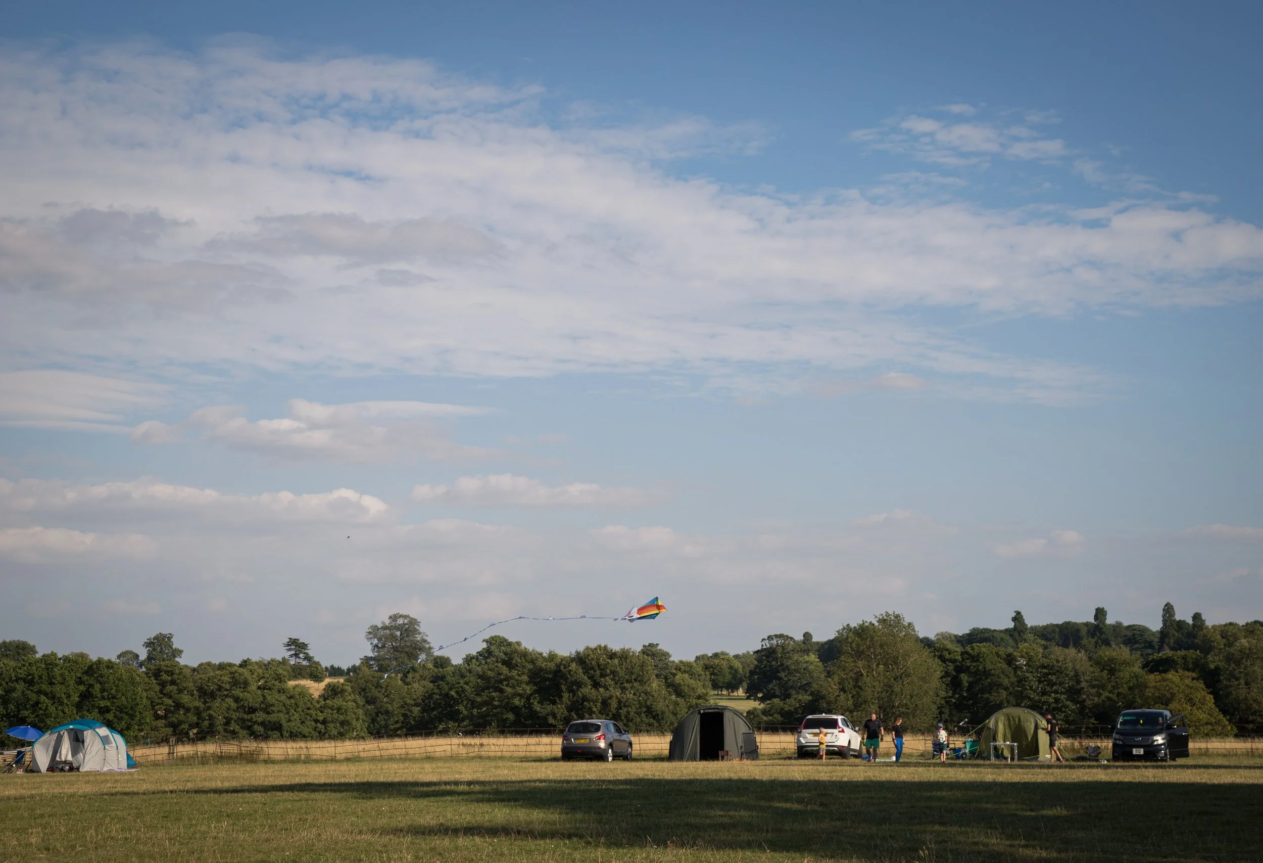 A group of people camping in a grassy field with tents and cars, flying a rainbow-colored kite under a blue sky with scattered clouds.