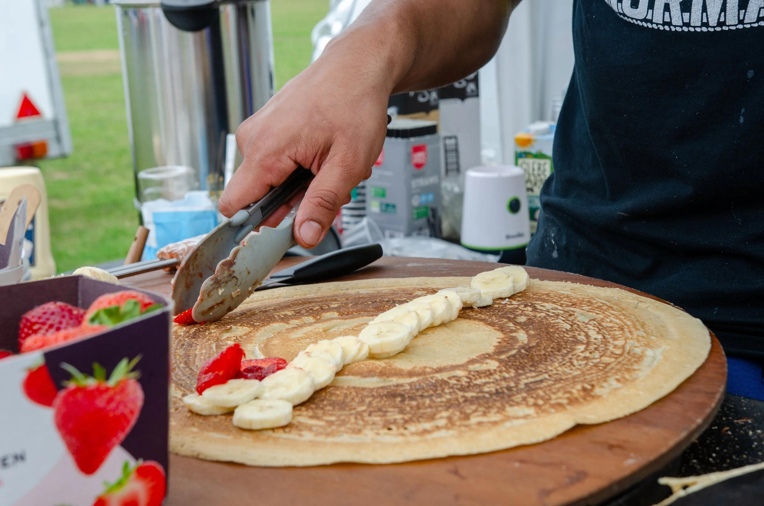 A person is making a crepe with sliced bananas and strawberries on a wooden surface, using tongs to place ingredients.