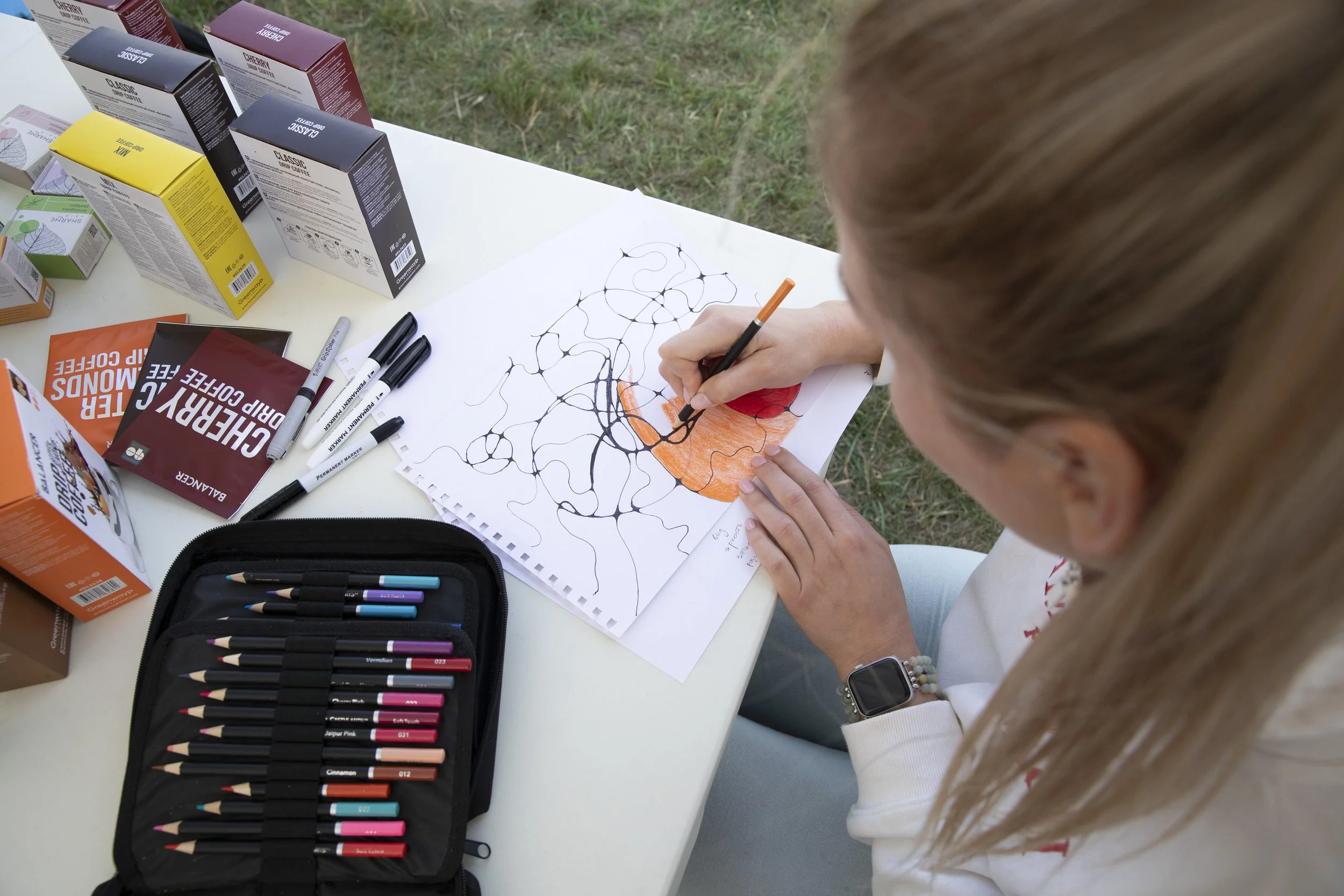 A young woman sitting at a white outdoor table, coloring on a piece of paper with black and orange markers. The table has various colored boxes of markers, some pens, and a black case with colorful markers. Grass is visible in the background.