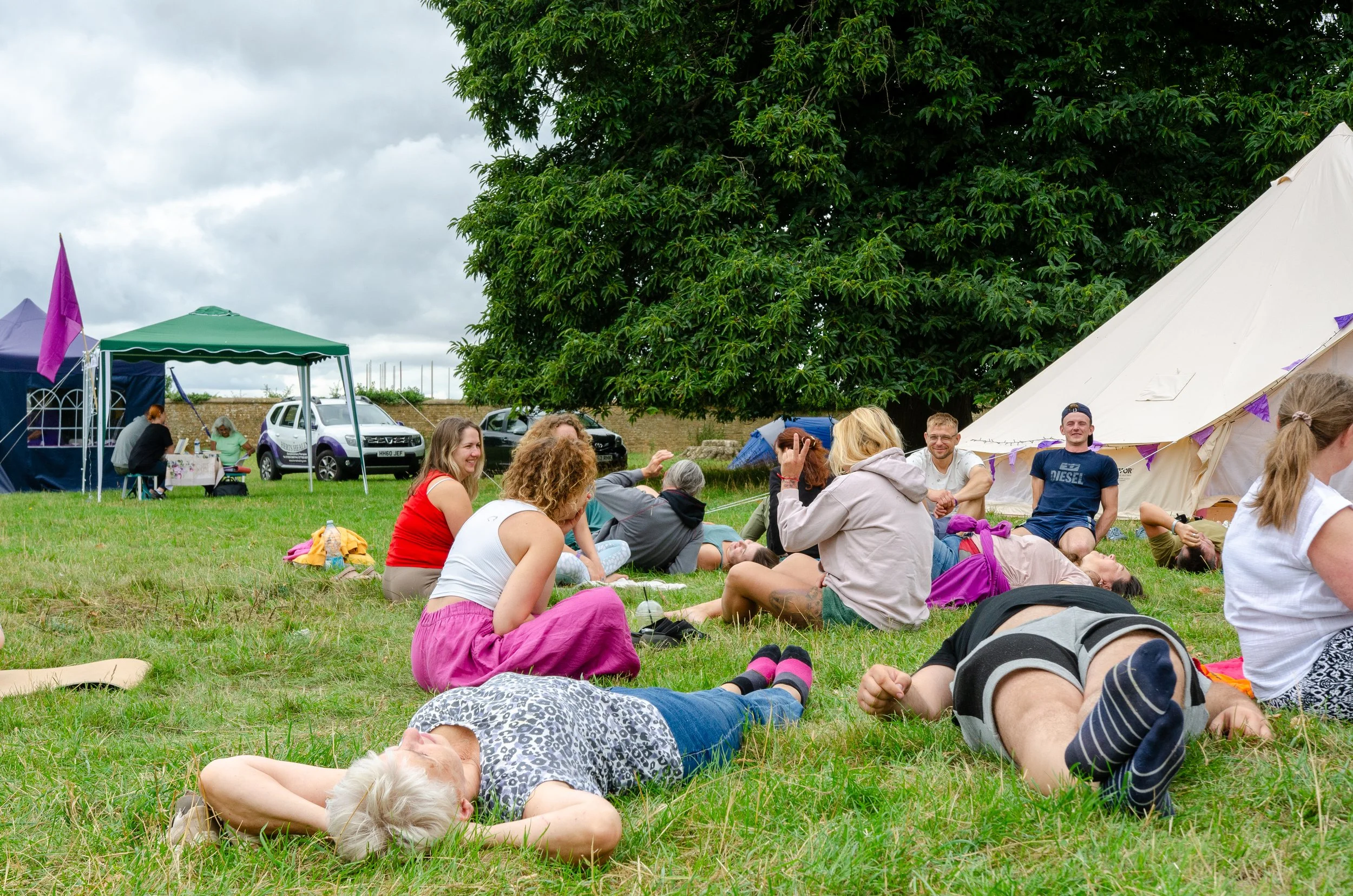 People relaxing on grass at an outdoor gathering with tents and trees in the background.