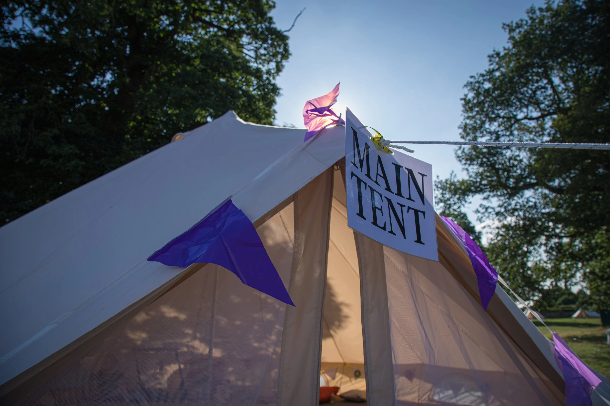 A white main tent with purple flag decorations and a sign hanging outside that reads 'MAIN TENT,' set against a background of trees and a blue sky.