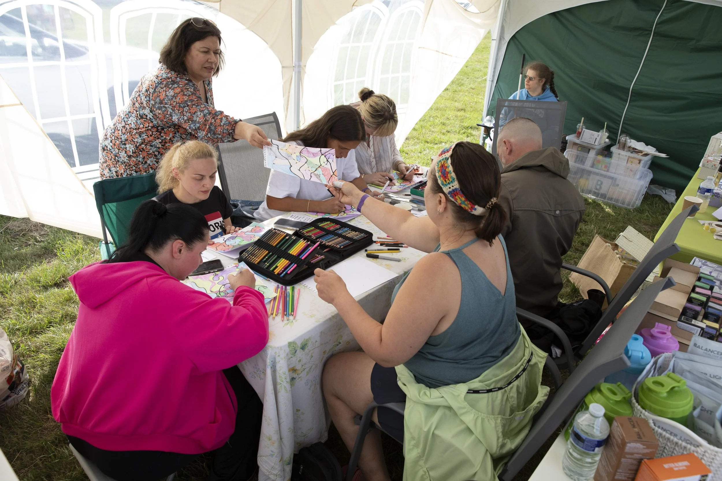 People sitting at a table inside a tent, engaging in arts and crafts, with art supplies like colored pencils and markers, and some are painting on paper.