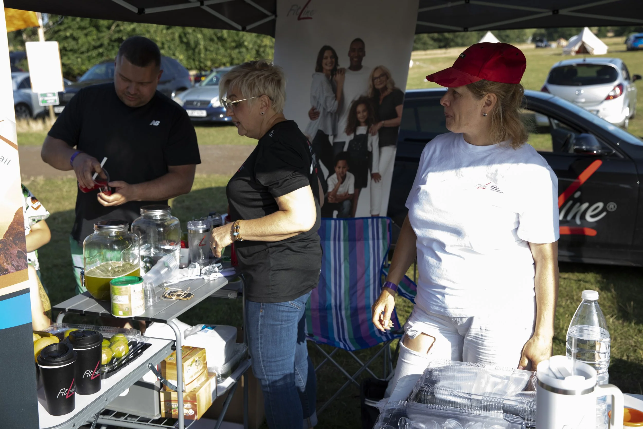Two women and one man at an outdoor event stand behind a table with drinks and supplies, with a car and a promotional banner in the background.