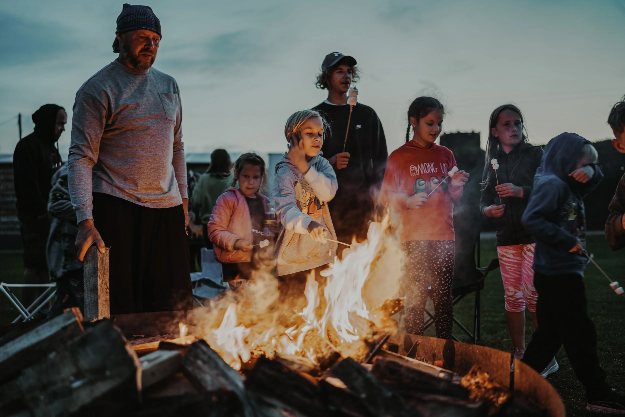 Group of children and adults gathered around a campfire at dusk, roasting marshmallows.