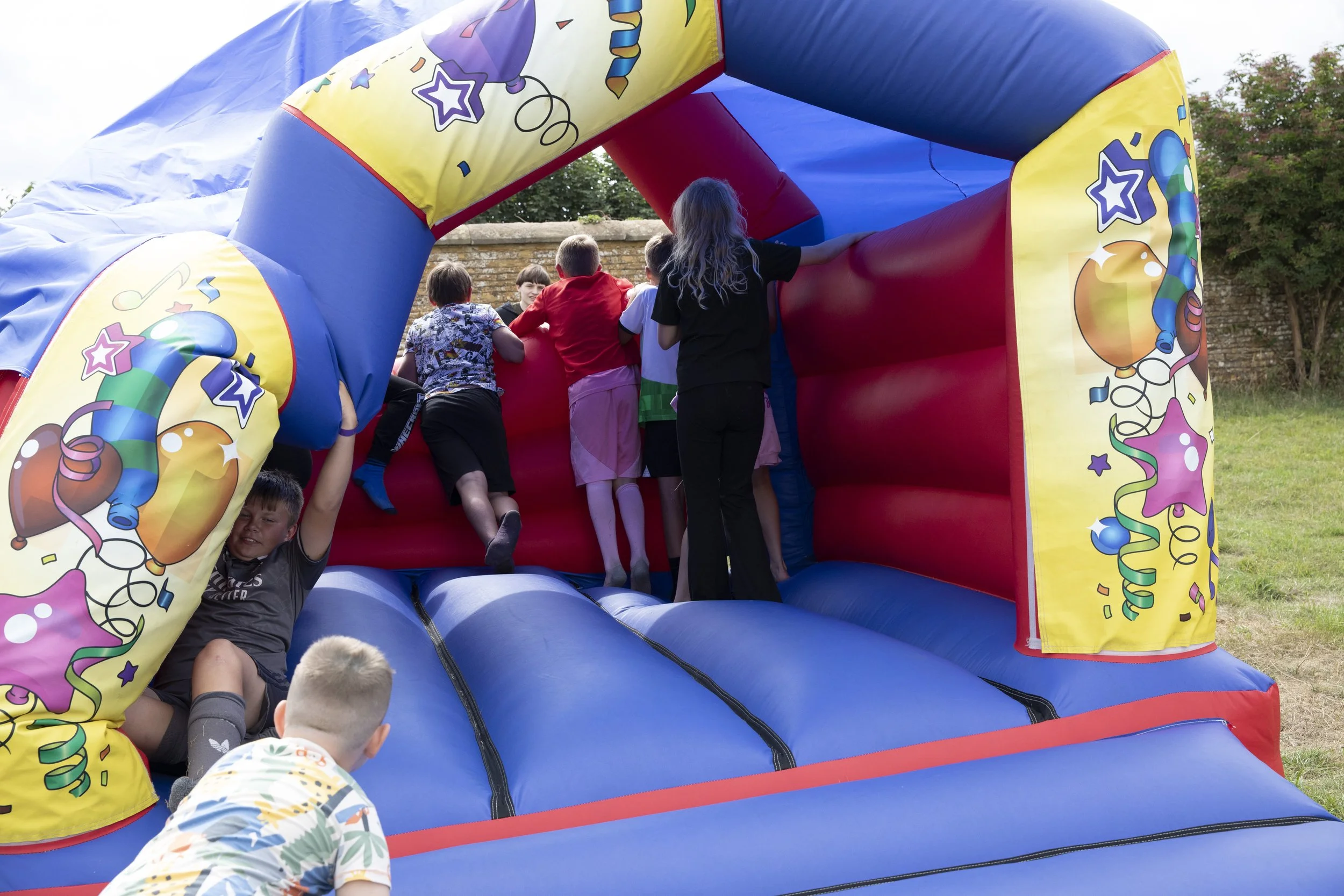 Children playing on a colorful inflatable bounce house with a party theme, decorated with balloons, stars, and streamers, outdoors on grass.
