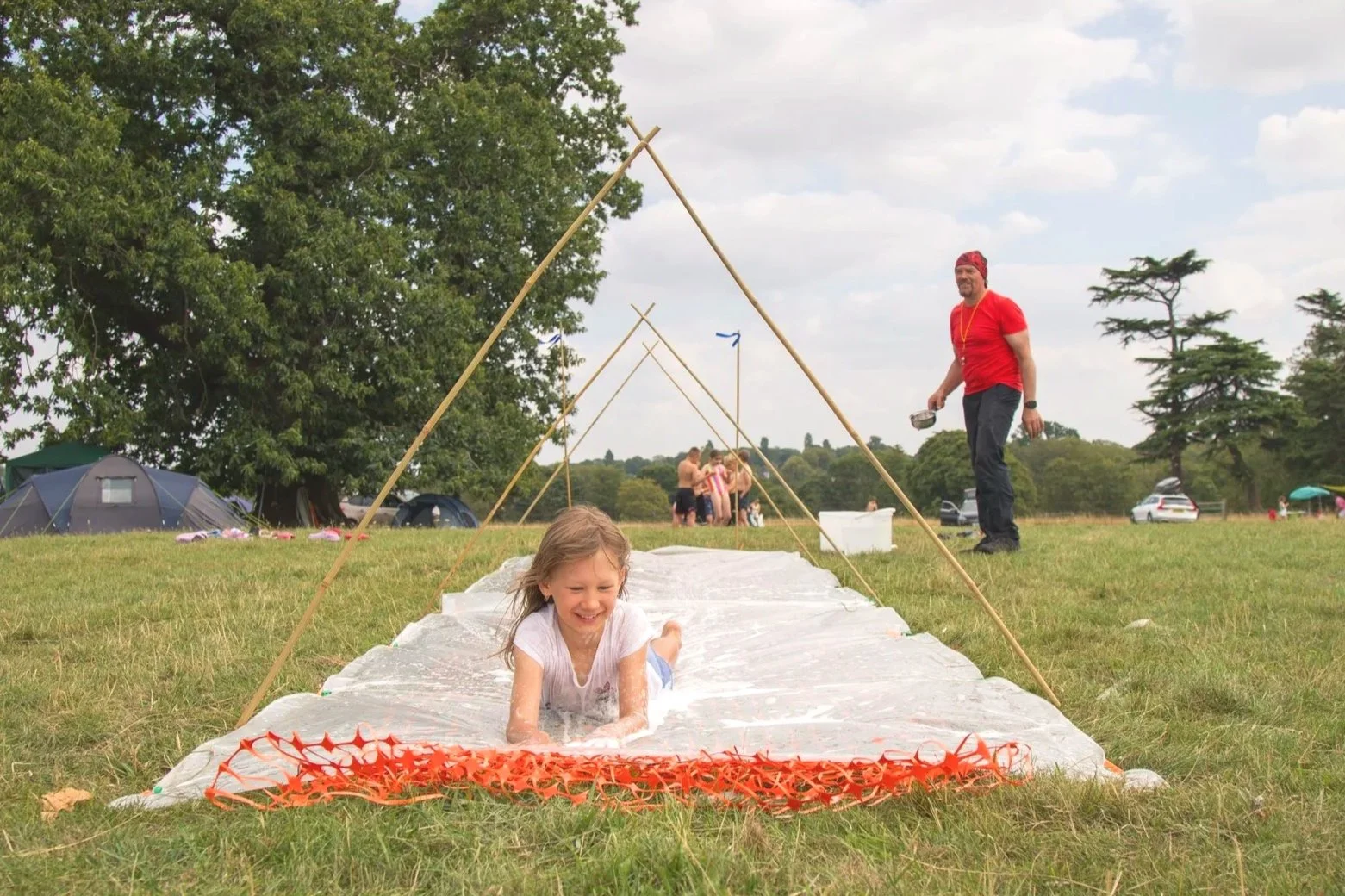 A young girl sliding down a makeshift slip-and-slide on a grassy field, with a man in a red shirt and red bandana supervising nearby during an outdoor event or camping trip.