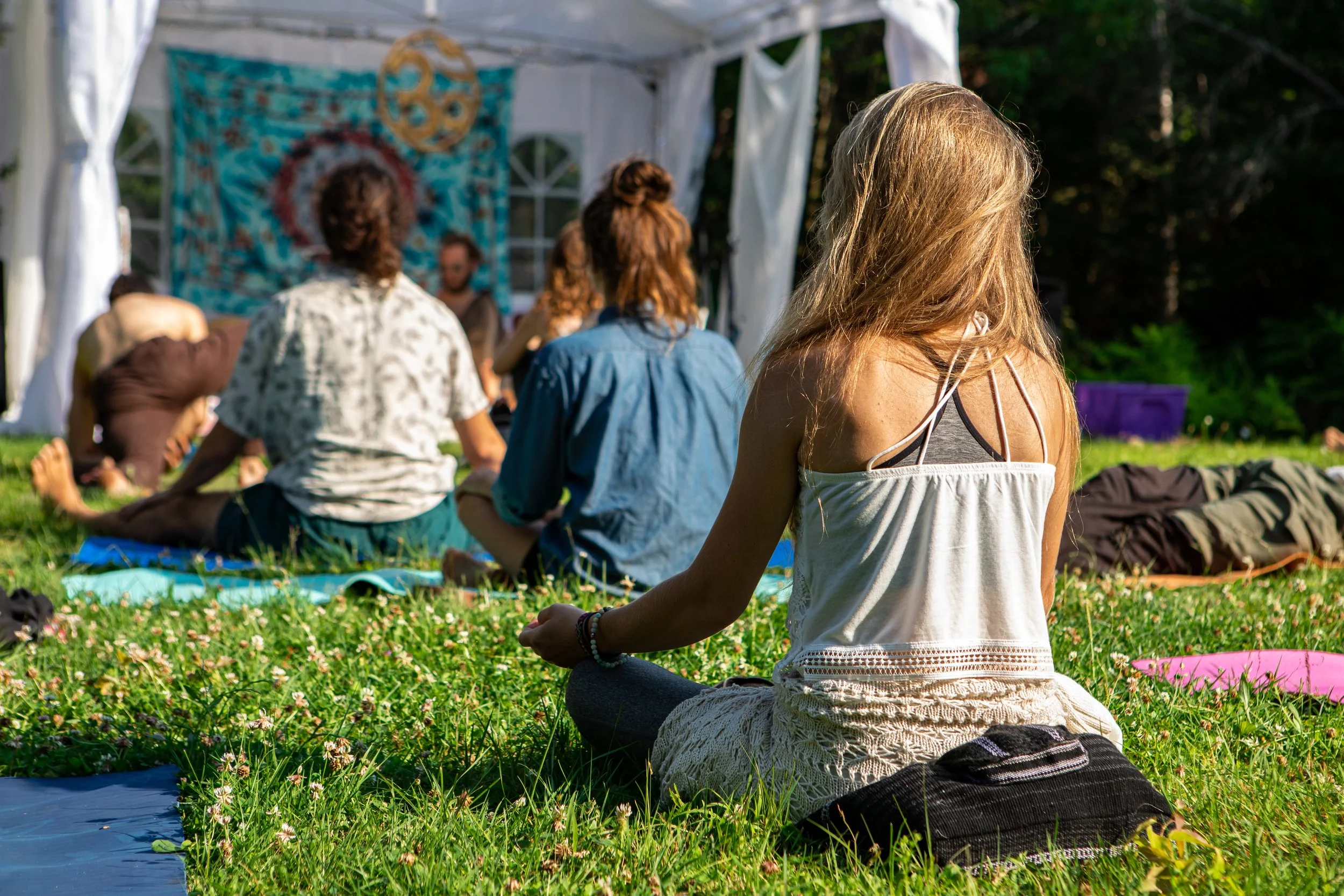 A group of people practicing yoga outdoors on grass near a white tent with a colorful tapestry, some in seated positions and one woman in the foreground sitting cross-legged with her back to the camera, wearing a white top with black straps.